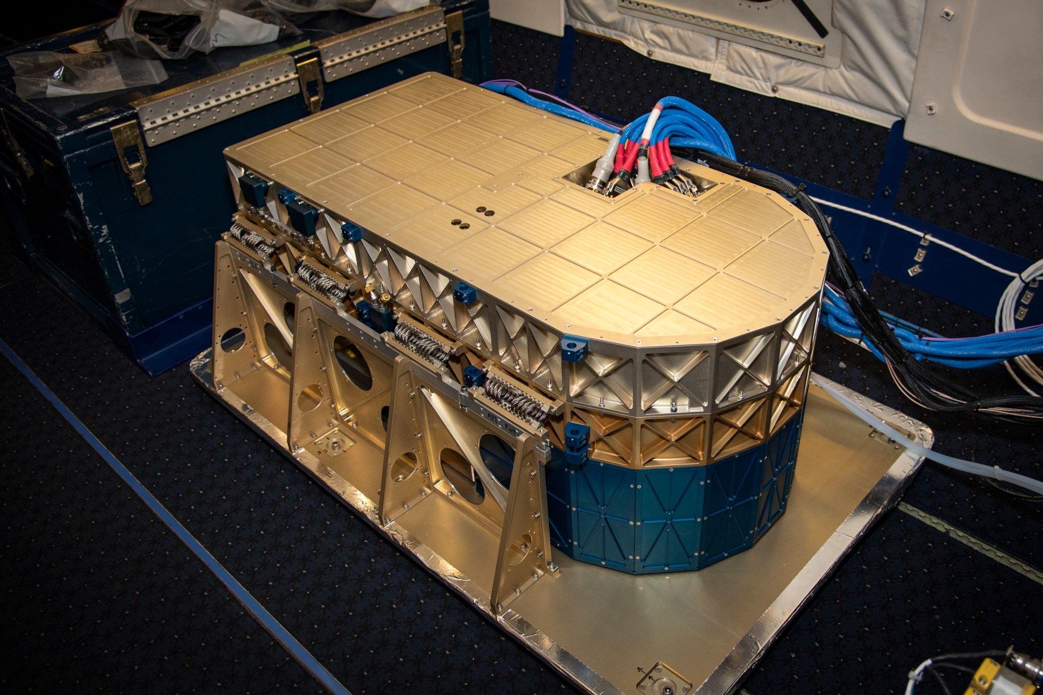 A gold instrument bolted to the floor of a research plane. It has three straight edges, with one curved edge. Wires poke out the mostly flat top.