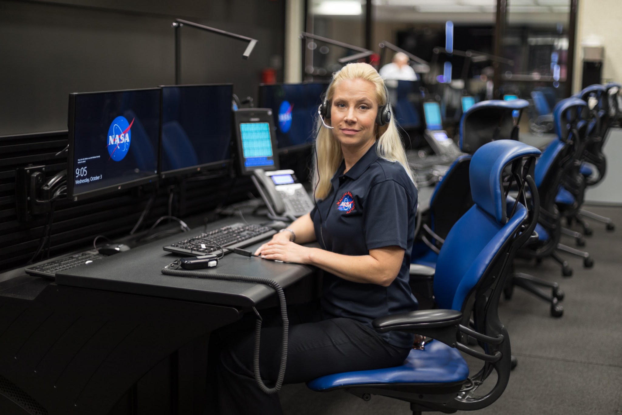 Woman with fair skin and long blonde hair looks at the camera while sitting in a blue chair in an operations console.