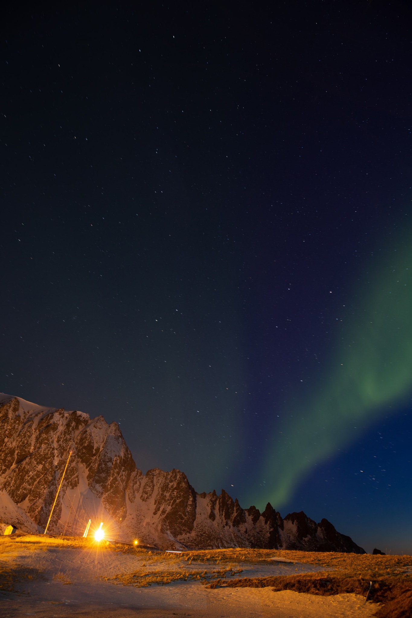 A bright green ribbon of light, the aurora borealis, glows against a dark blue night sky with a few visible stars. The streams of light seem to extend from the far distance. In the foreground, reddish dirt ground leads to a range of jagged brown mountains. On the right, a small collection of white buildings are visible.