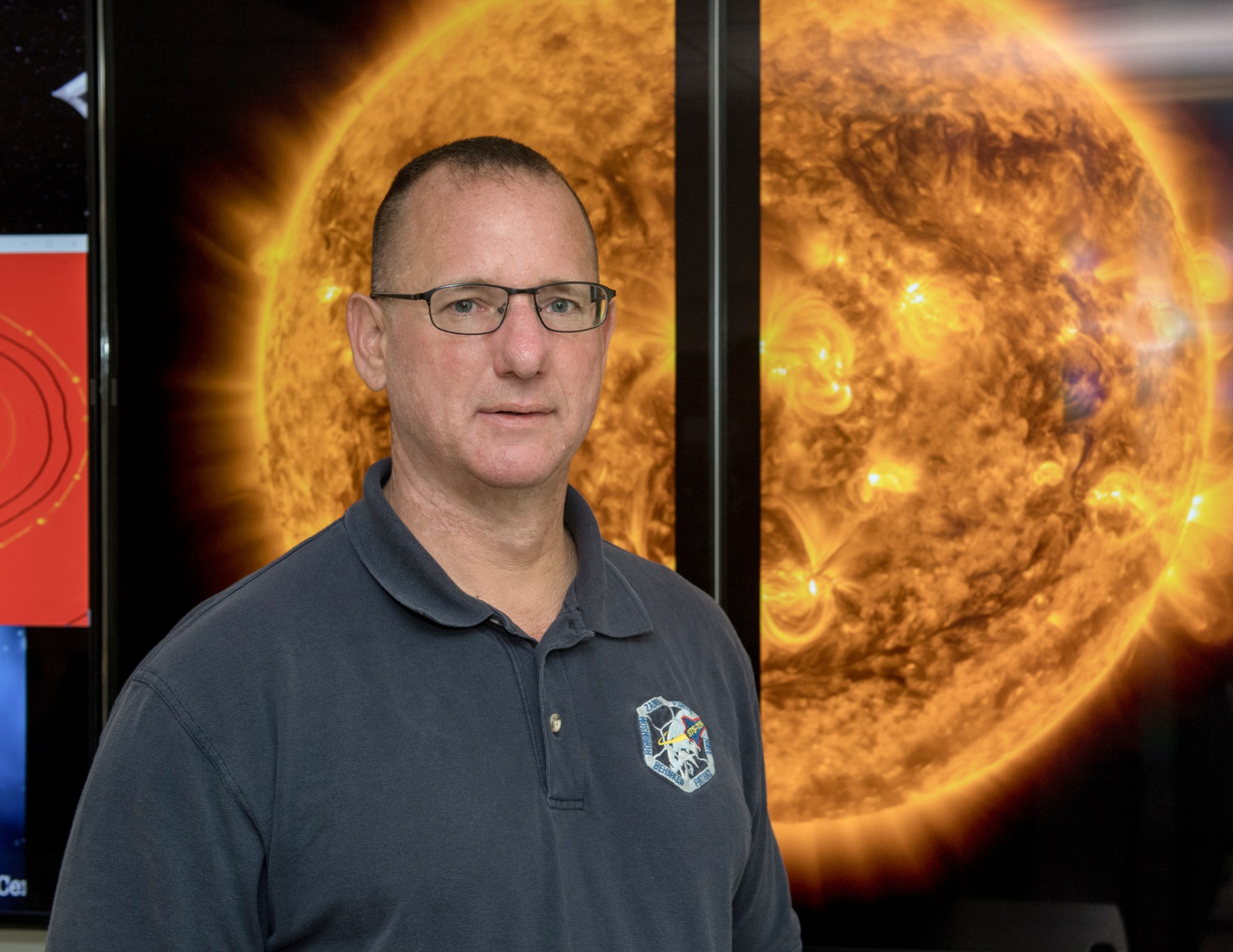 Man with fair skin and brown hair wears glasses and a grey mission polo. He is standing in front of a hyperwall image of the Sun. 
