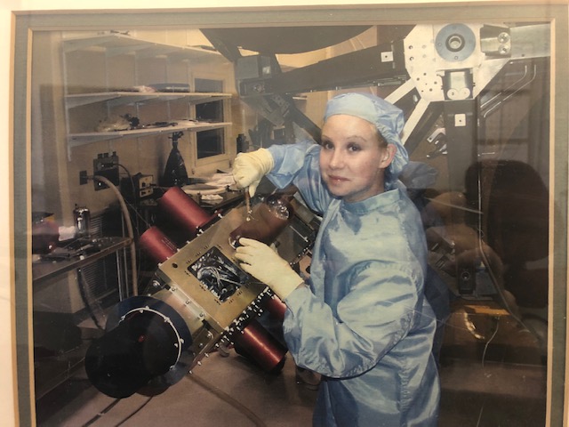 Older photo of woman wearing a blue lab coat, white gloves, and a blue hair cover and is working on a control module