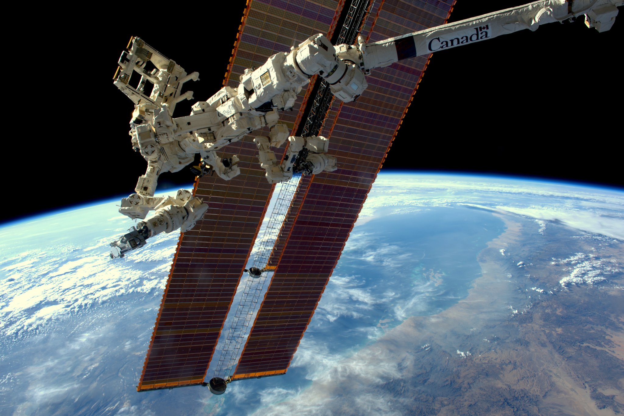 RELL in orbit at ISS. The instrument is white and held at the end of a mechanical arm. Behind the arm, two dark gold solar panels float above the light blue limb of Earth.