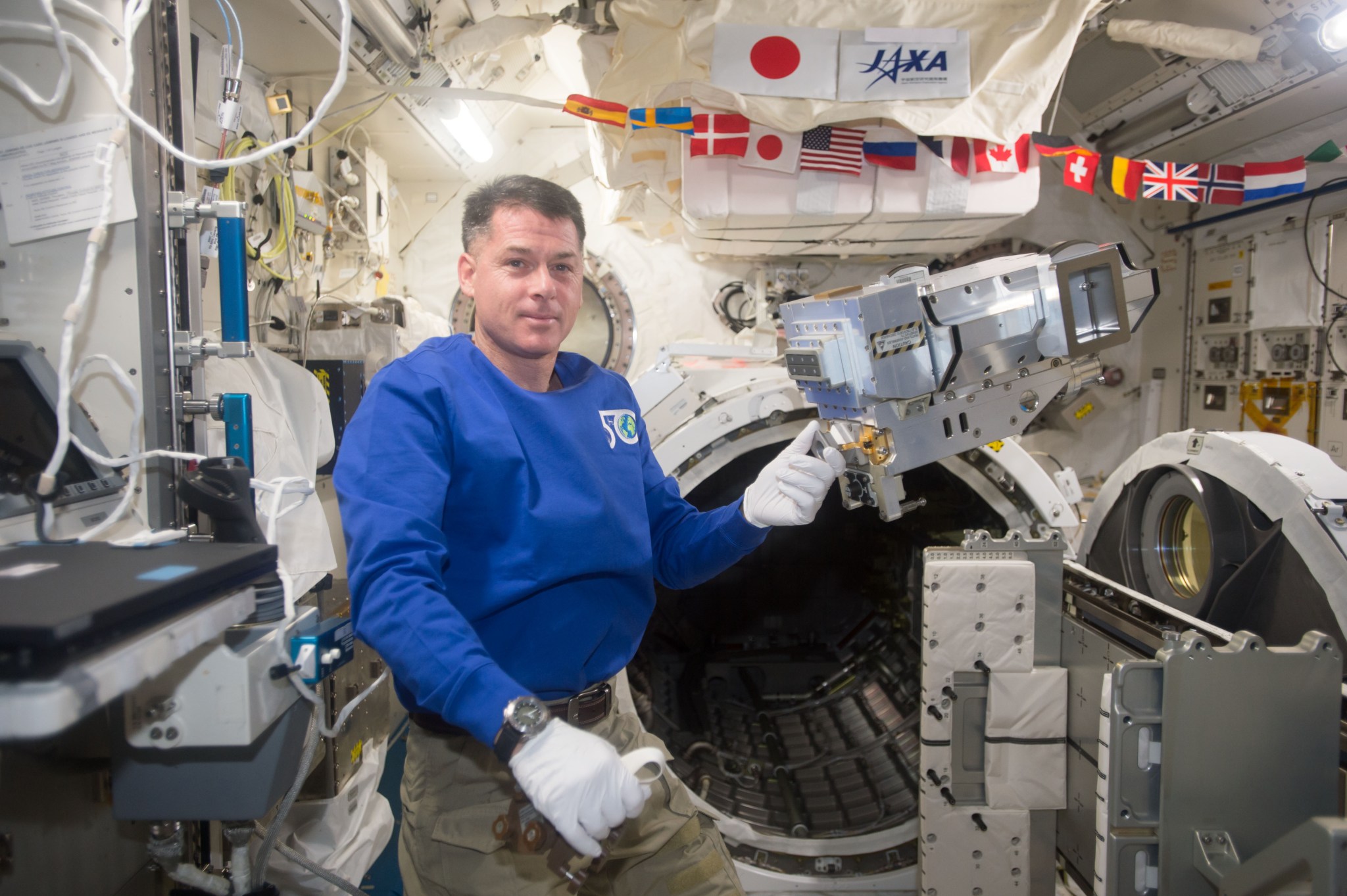 Shane Kimbrough and leak locator on the Space Station. He is wearing a blue shirt and white gloves and gesturing to the leak locator, which is floating in microgravity.