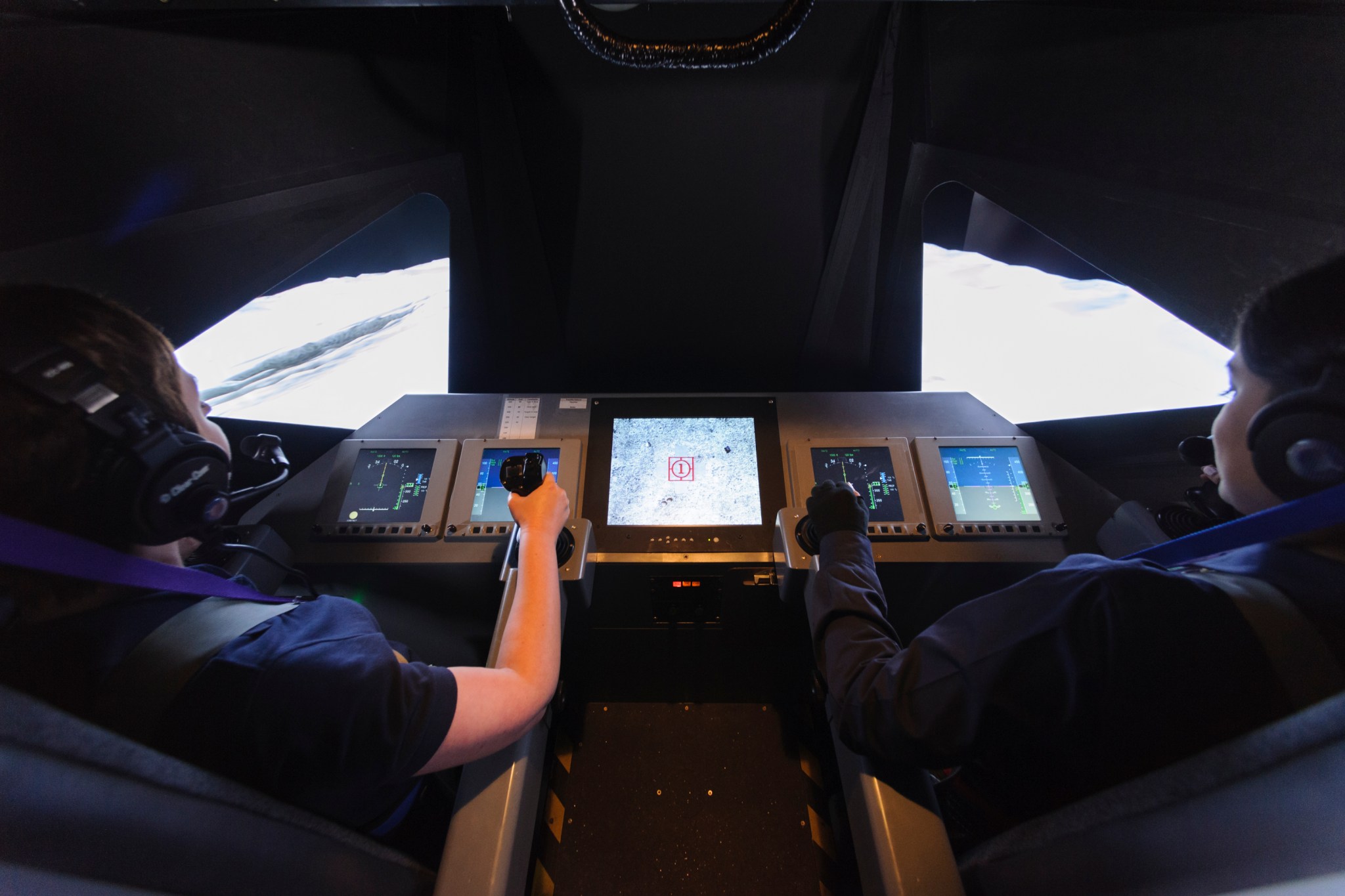 Two pilots in a mock lunar lander cockpit. Realistic flight controls are seen and graphics of the lunar surface out the window.