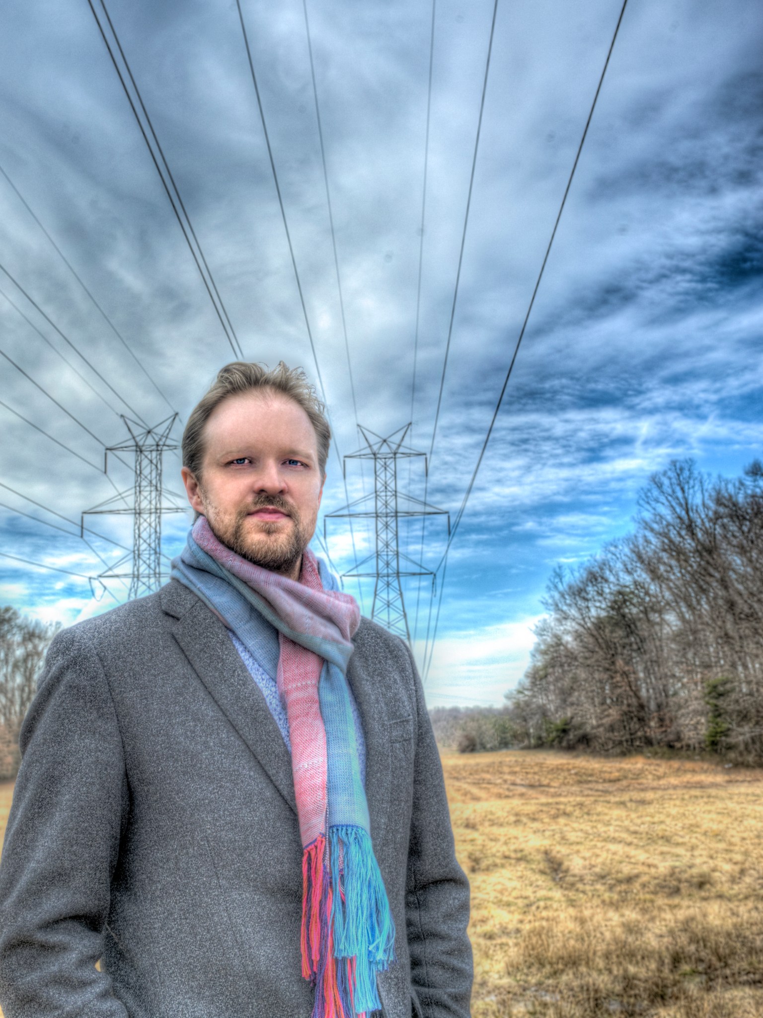 A man stands underneath power lines. The man has brown hair and brown facial hair. He is wearing a blue and pink scarf and a gray coat. 