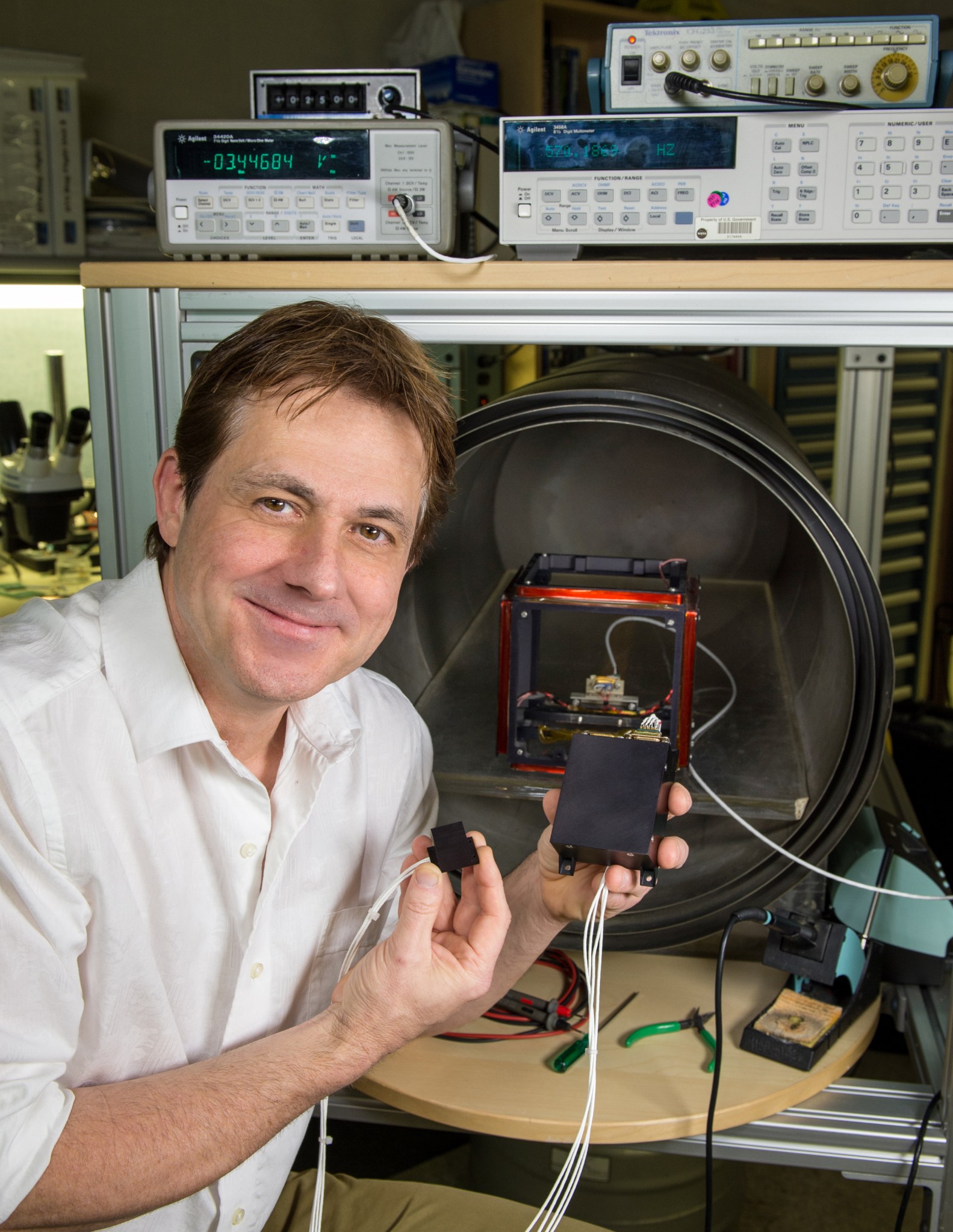 Man with brown hair and fair skin smiles holding a small black box in one hand and a smaller black box in the other, they are attached by several white cords. 