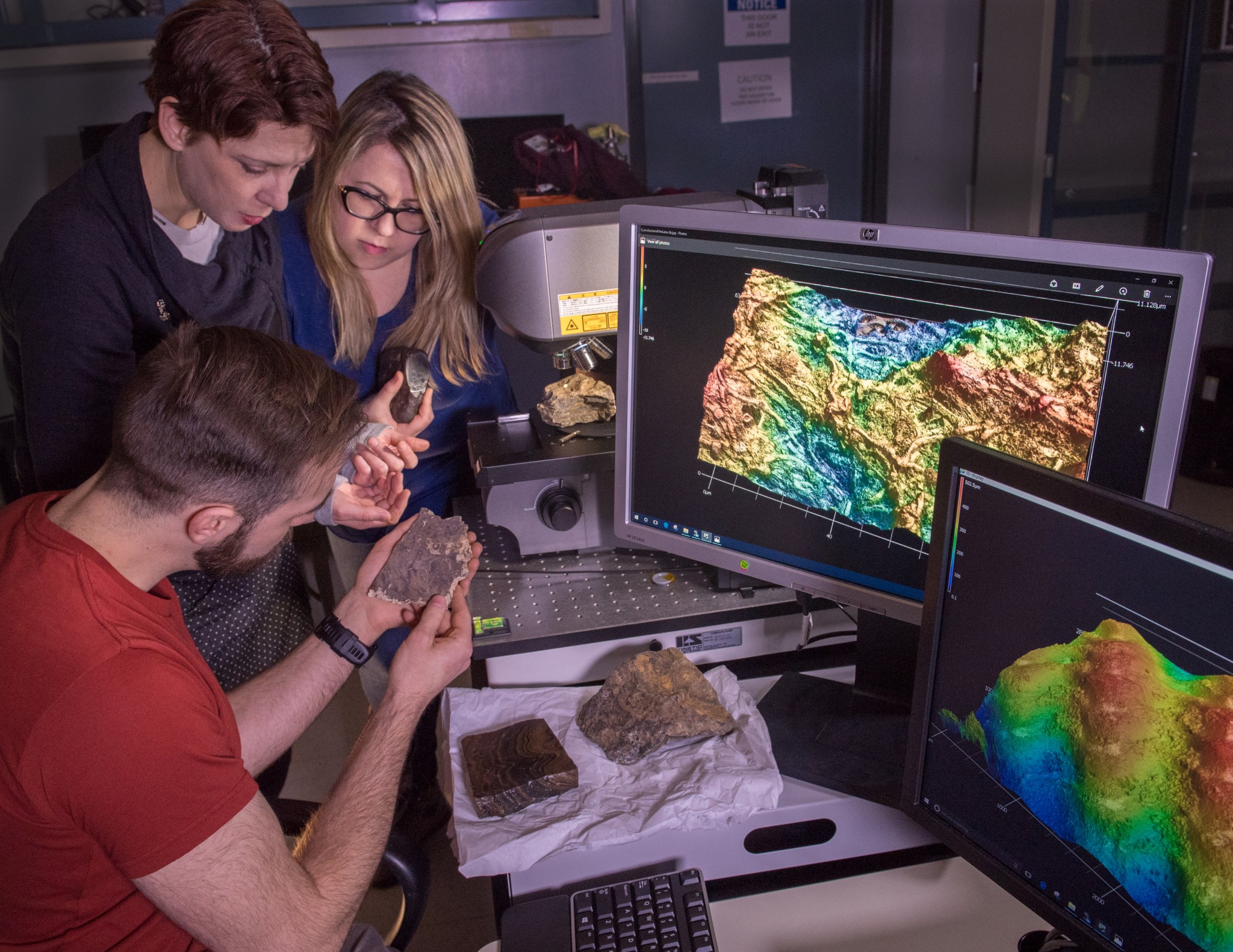 Three people examine rock and wood samples as a topographical map is shown on the screens in front of them.