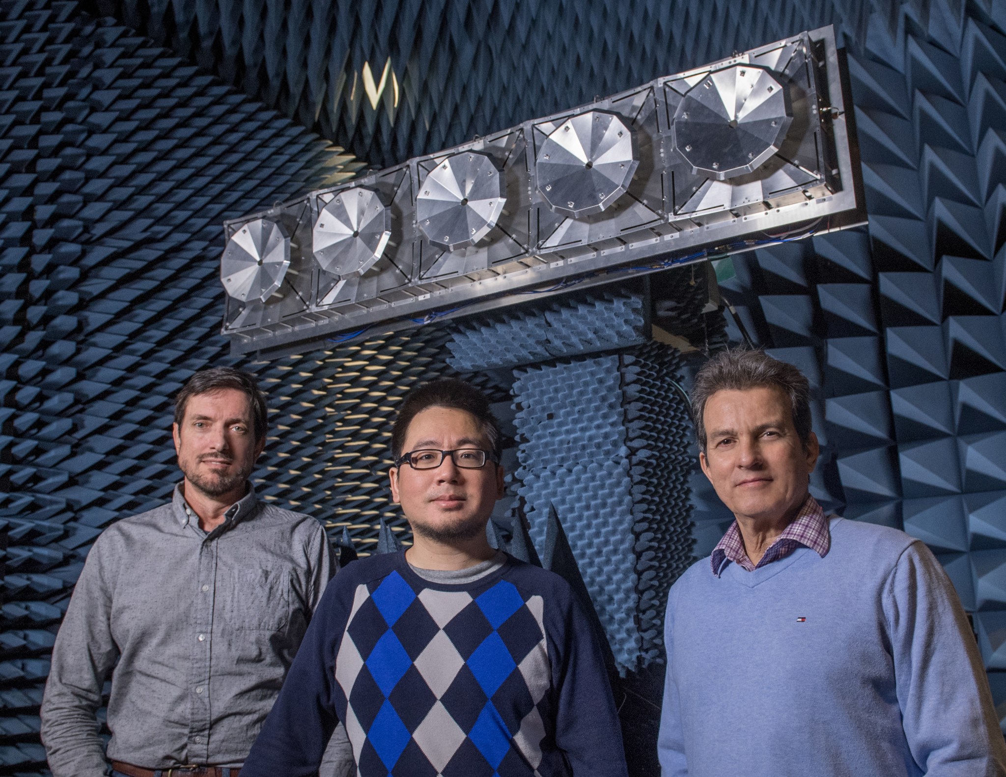 Three men stand in a prototype antenna subarray for Space Exploration Synthetic Aperture Radar.  The background looks like hundreds of small foam pyramids and there is a silver panel of five circles at the top. 