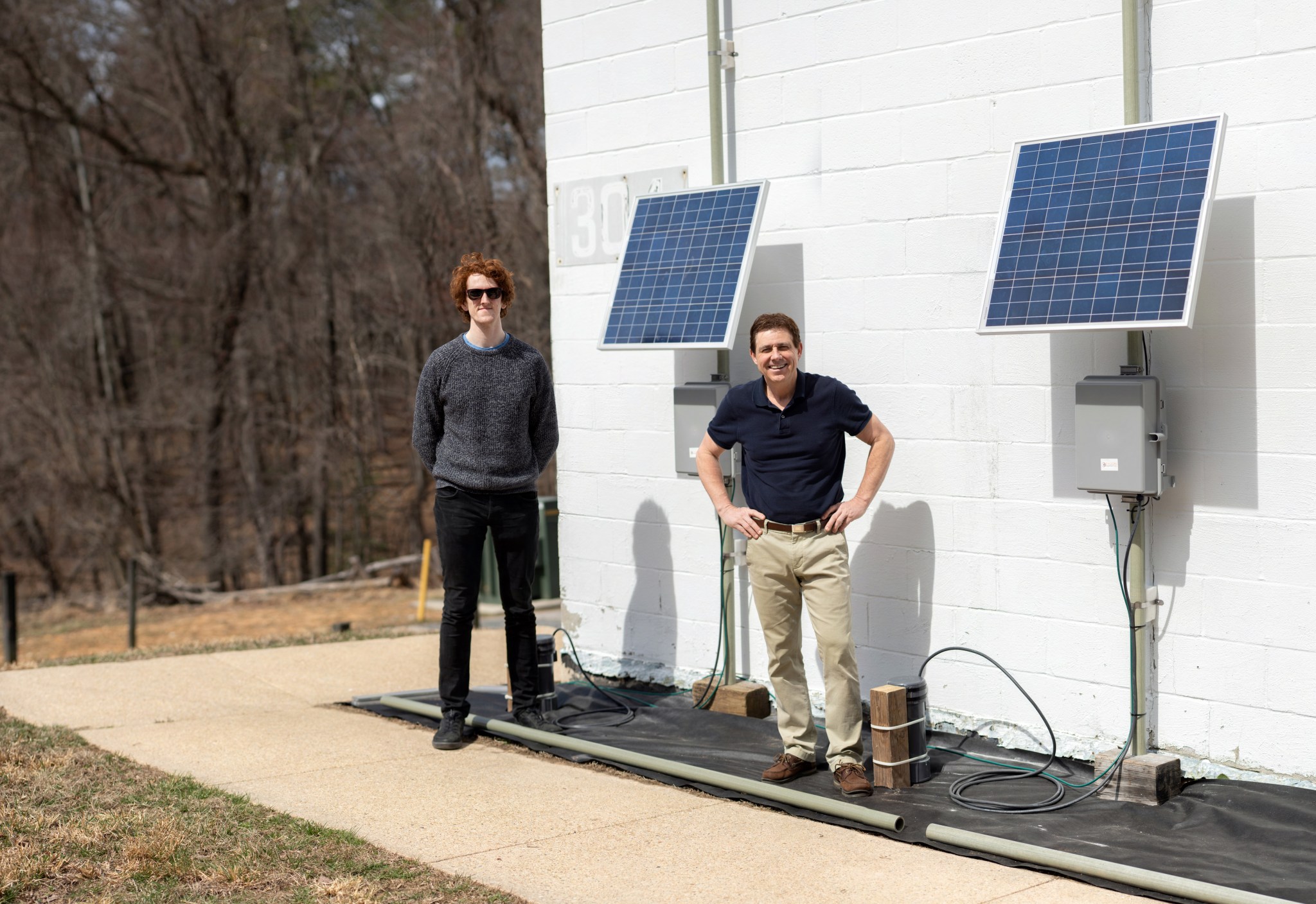 Two men stand next to power lines looking at the camera. There re two panels connected to boxes and lines on the wall behind them. 