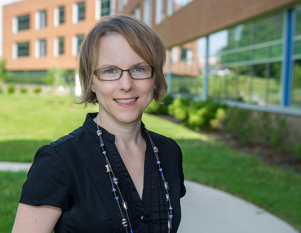 A woman with short hair and glasses smiles with a building in the background