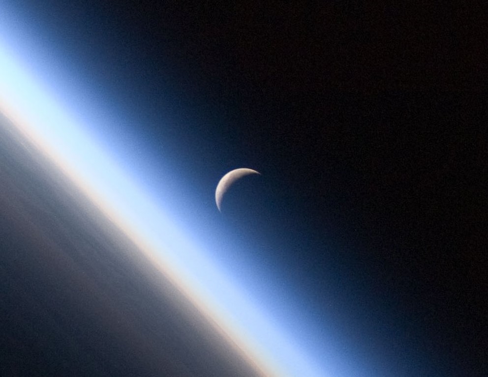 A setting, waning crescent moon amid the thin line of Earth's atmosphere.