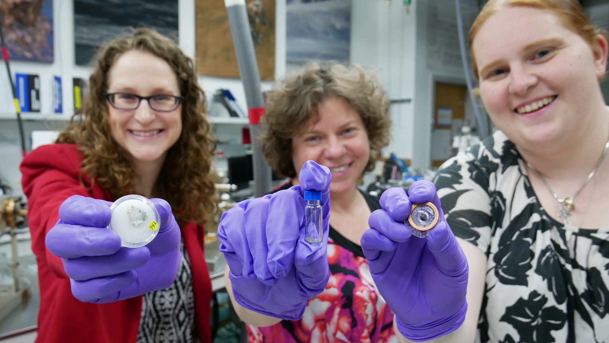 Three female scientists are smiling at the camera and holding Apollo samples. 