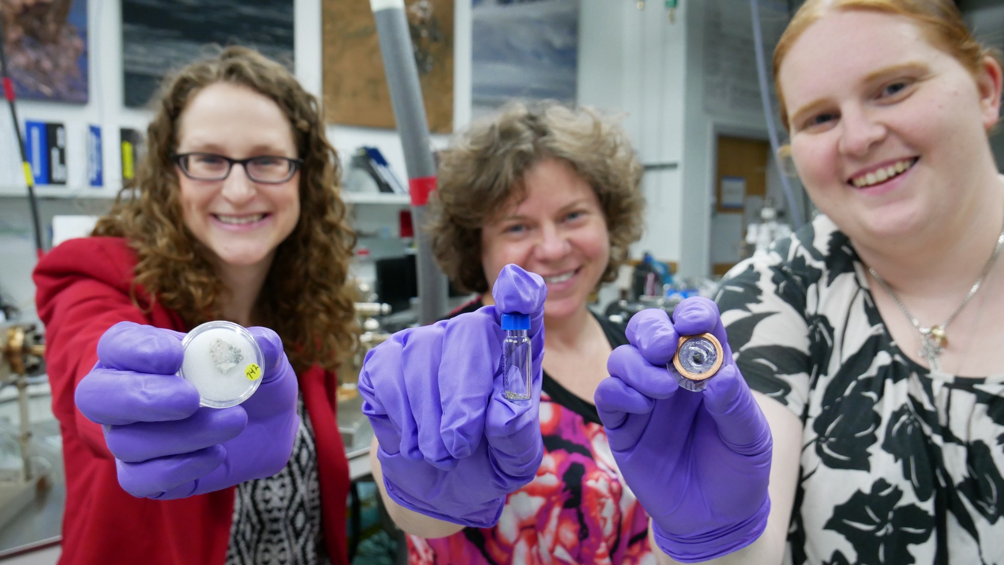 Three female scientists are smiling at the camera and holding Apollo samples. 