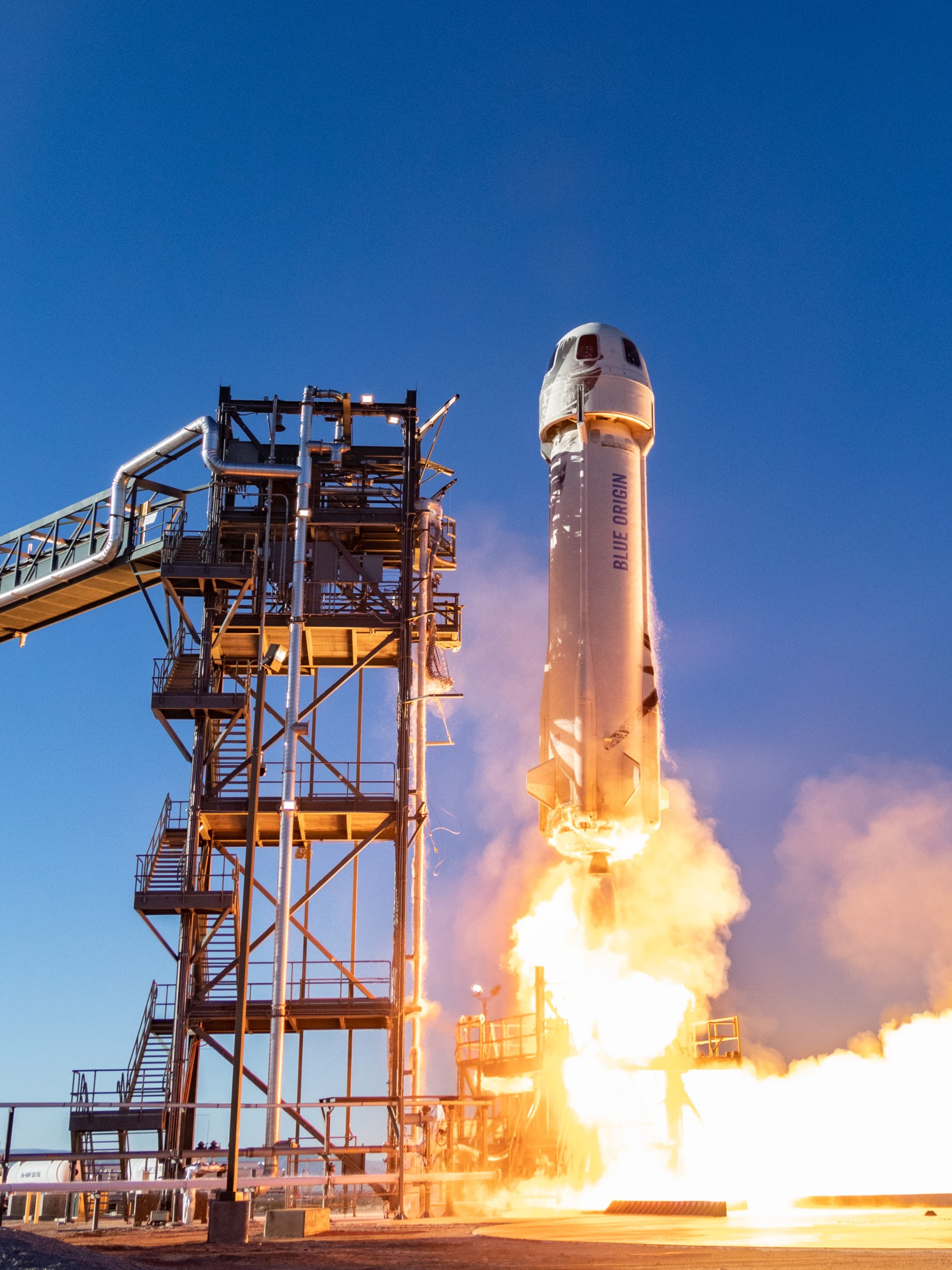 Rocket with "Blue Origin" on the side lifting off on the launch pad. There is fire and smoke coming from the bottom of the rocket as it lifts off. 