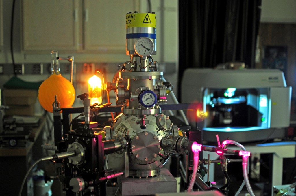 A laboratory showing equipment from the In situ Carbon Evolution Experiments (ICEE) chamber. Bright yellow flask and a glowing substance coming from one tube featuring high power pressure valves that have bright pink colors coming from the tubing.