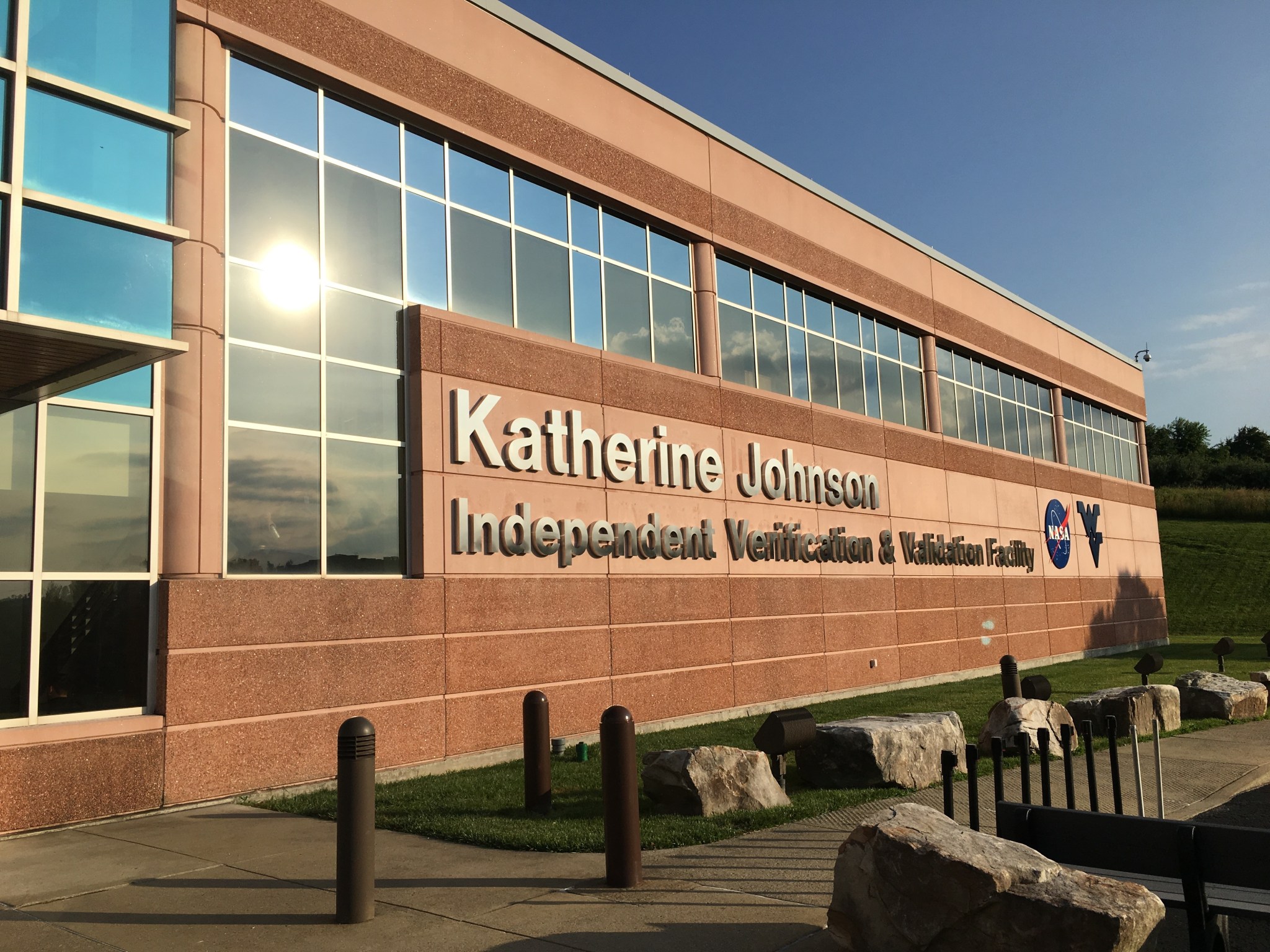 The side of a brown brick building with windows glistening in the sunlight. The words Katherine Johnson Independent Verification and Validation Facility with the NASA meatball logo and a WV for West Virginia.