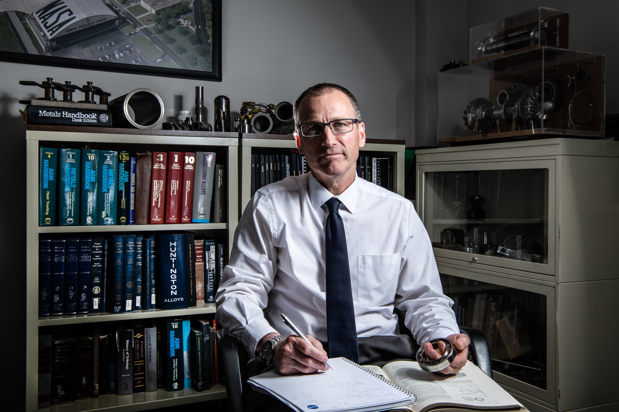 Dr. Christopher DellaCorte wears a white shirt, tie, and glasses, is shown facing the camera as he sits in an office with book shelves visible behind him. He holds an open notebook, a pen, and Ball bearings made of Nitinol shape-memory alloy won’t rust or corrode.  
