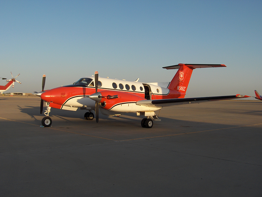 The King Air plane sitting on a runway. It's a small plane with just six windows on the side, painted red and white with a dark stripe.
