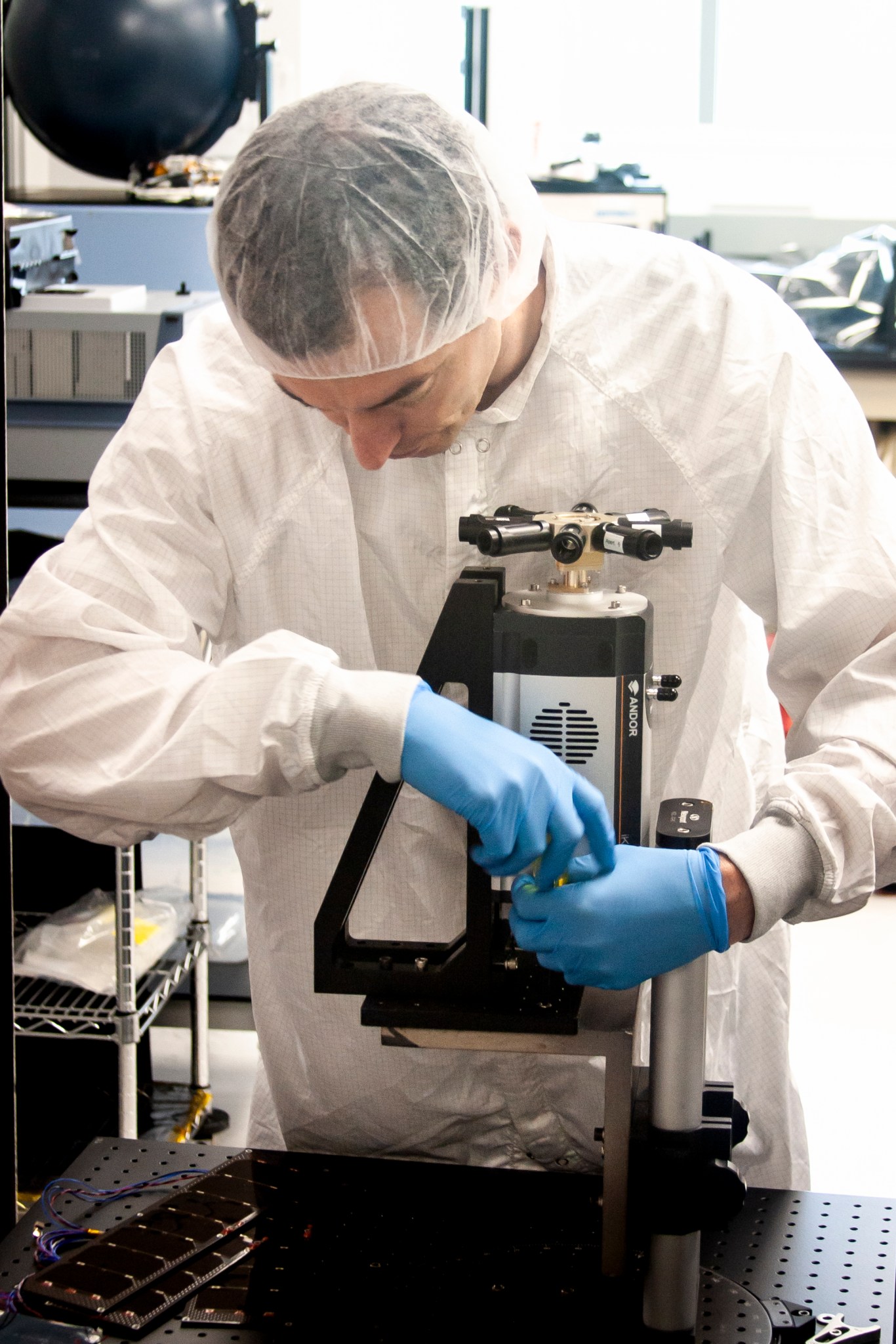 Man with fair skin and brown hair wears a lab coat, hair net, and blue gloves and operates an instrument on a table, using both hands