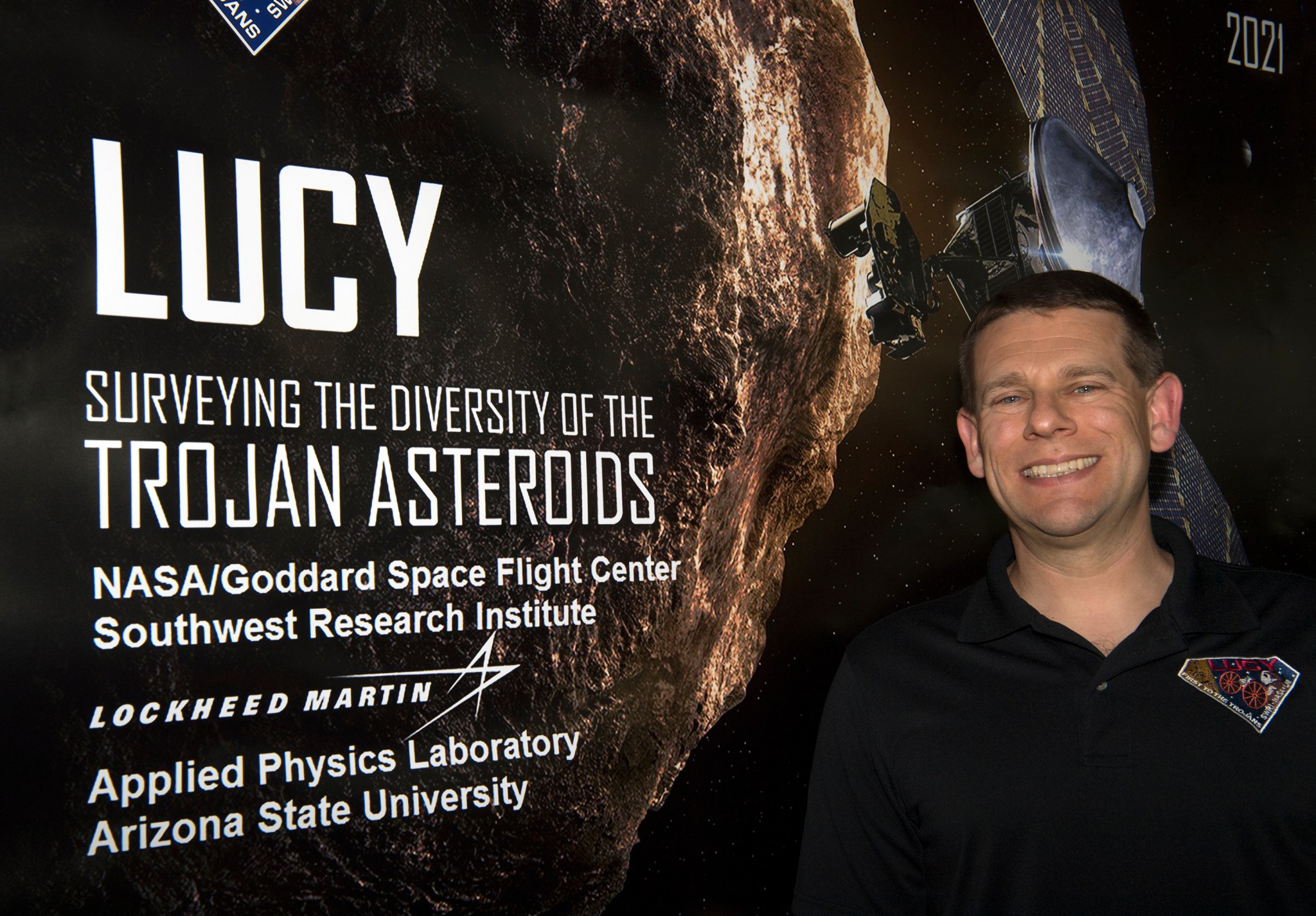 Man with brown hair and fair skin stands smiling next to a poster for the Lucy mission