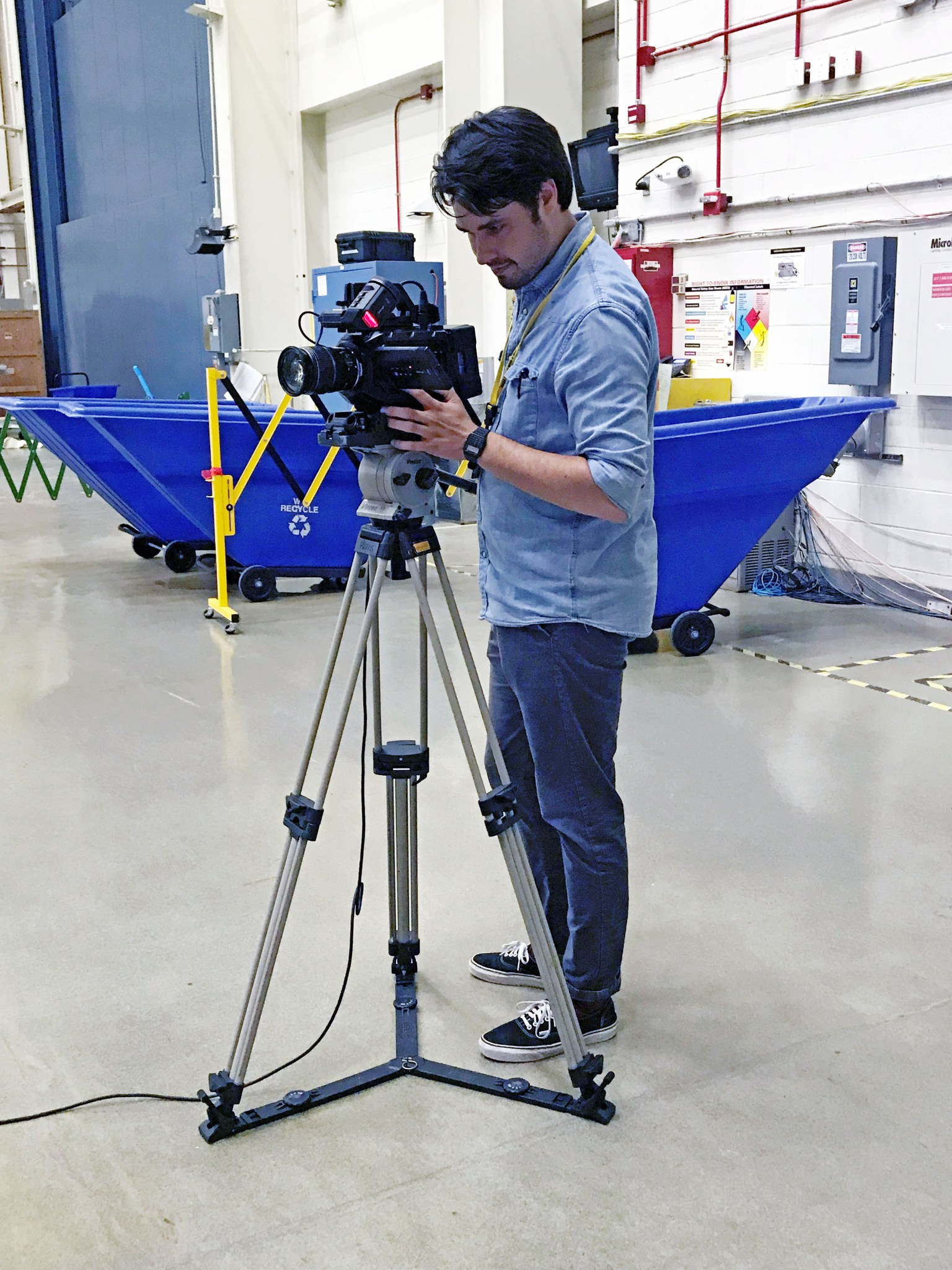 Man wearing a blue button down with jeans and vans looks left operating a video camera on a tripod. There are large blue recycling carts behind him. 