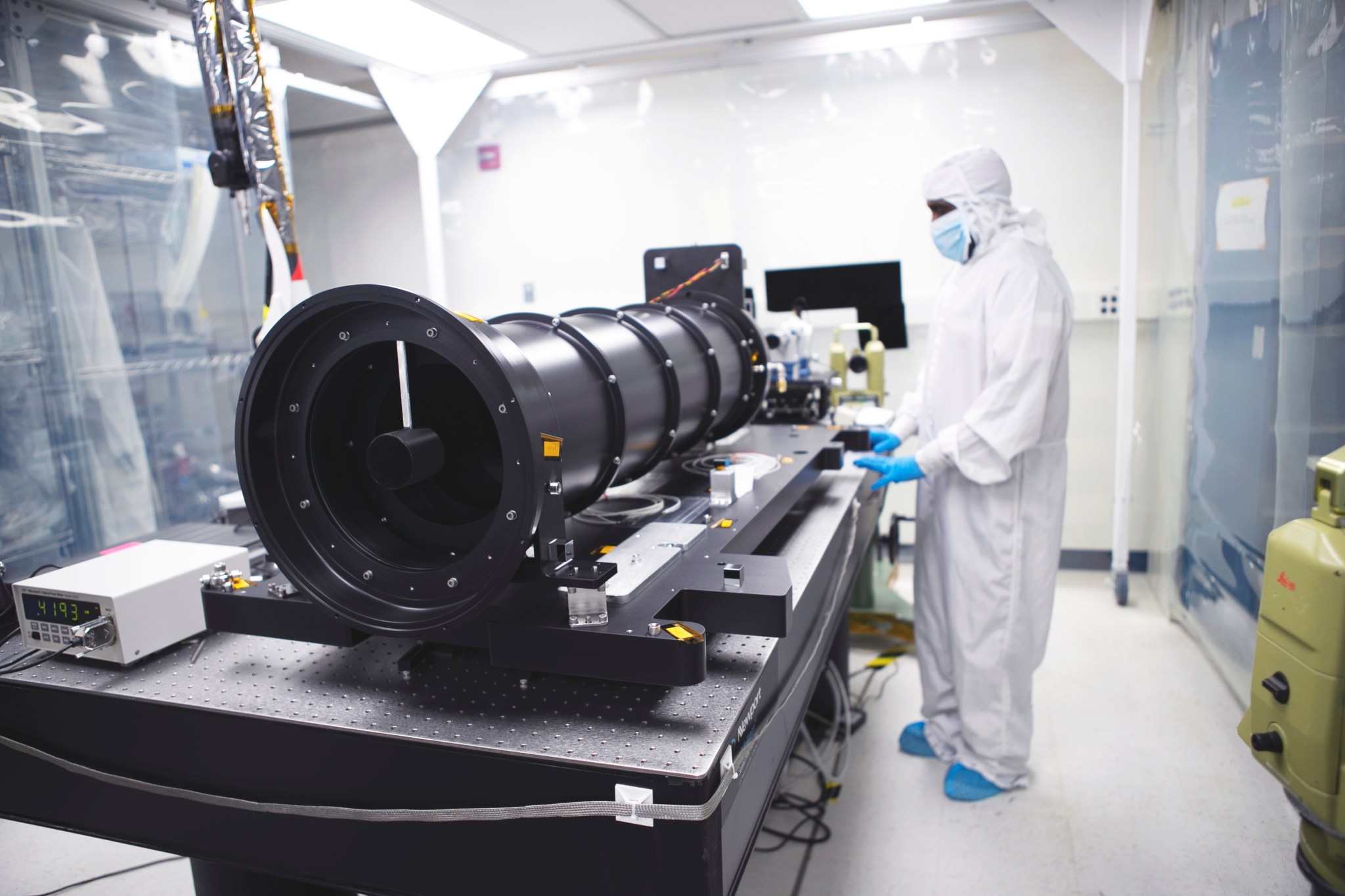 A person in a white suit stands next to a coronagraph instrument, which appears as a long, black tube, in clean room with white walls and floors.