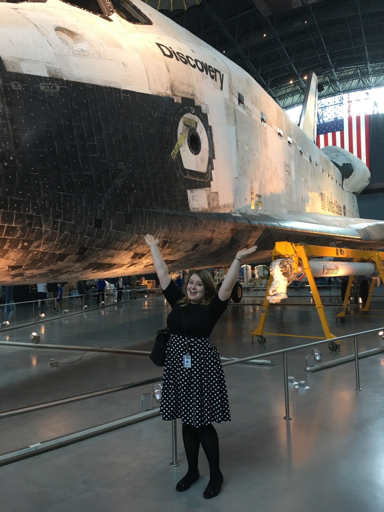 woman with shoulder length brown blonde hair wears a black and white polka dot skirt with a short sleeved black shirt has her arms up and is smiling in front of the Space Shuttle, "Discovery"