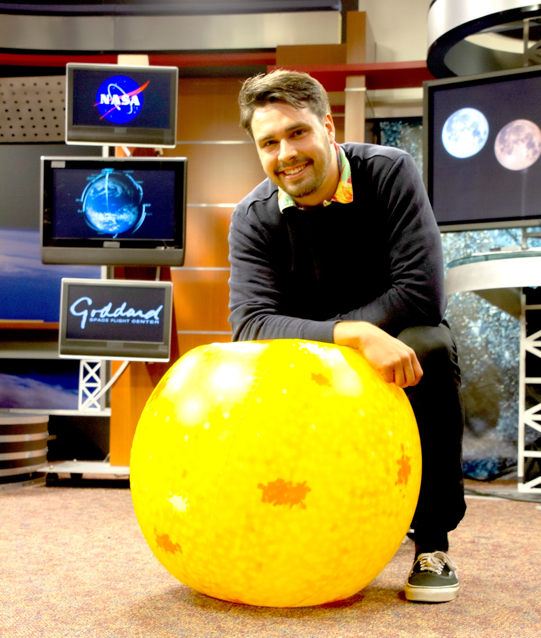 Harrison Bach kneels next to an inflatable Sun in the Goddard tv studio. He is wearing a black sweater with a  multicolored collared shirt under, black pants, and black vans. 