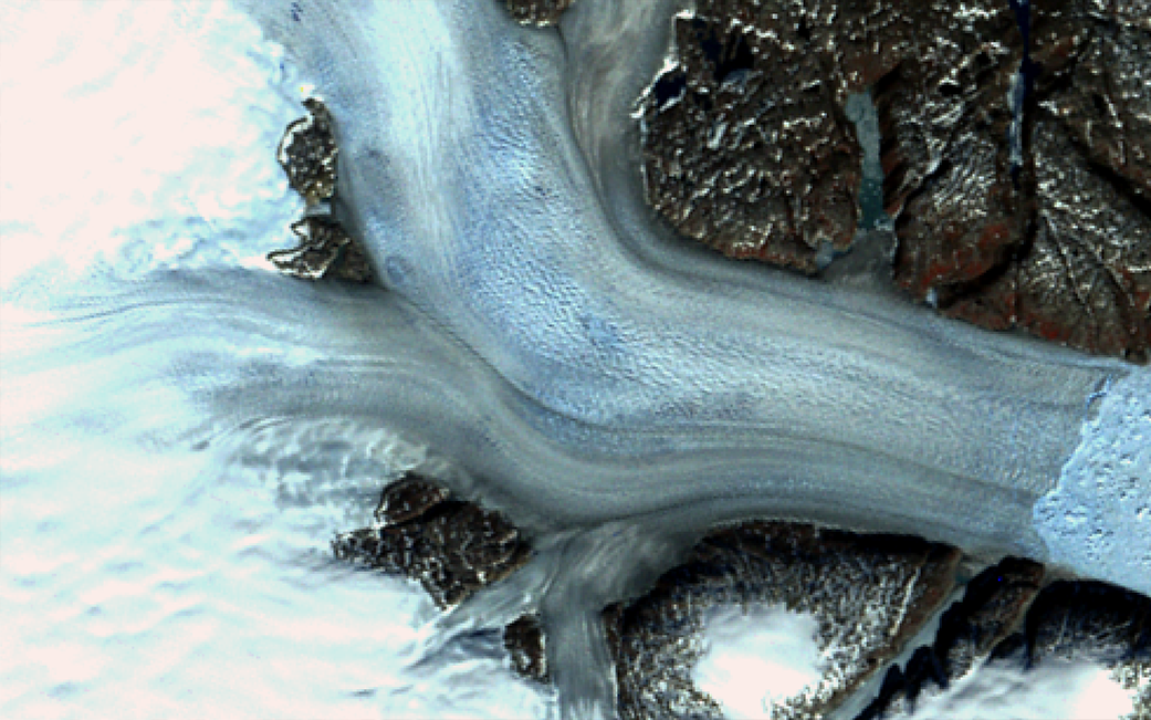 Satellite image of Helheim Glacier flowing like a river to the sea. The age of the image means it's in false color, so the ice looks bright blue, and the surrounding land looks reddish.