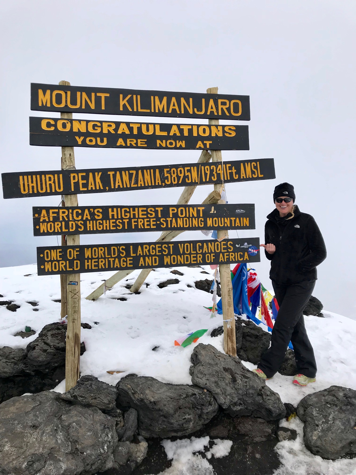 Woman with light skin wearing a black jacket, black pants, green and pink tennis shoes, a black beanie, and sunglasses stands on snow-covered rocks, smiling and pointing at a sign with the words "Mount Kilimanjaro, Congratulations you are now at, Uhuru Peak, Tanzania, 5895M/19341 ft AMSL, Africa's highest point, World's highest free-standing mountain, one of the largest volcanos, world heritage, and wonder of Africa.