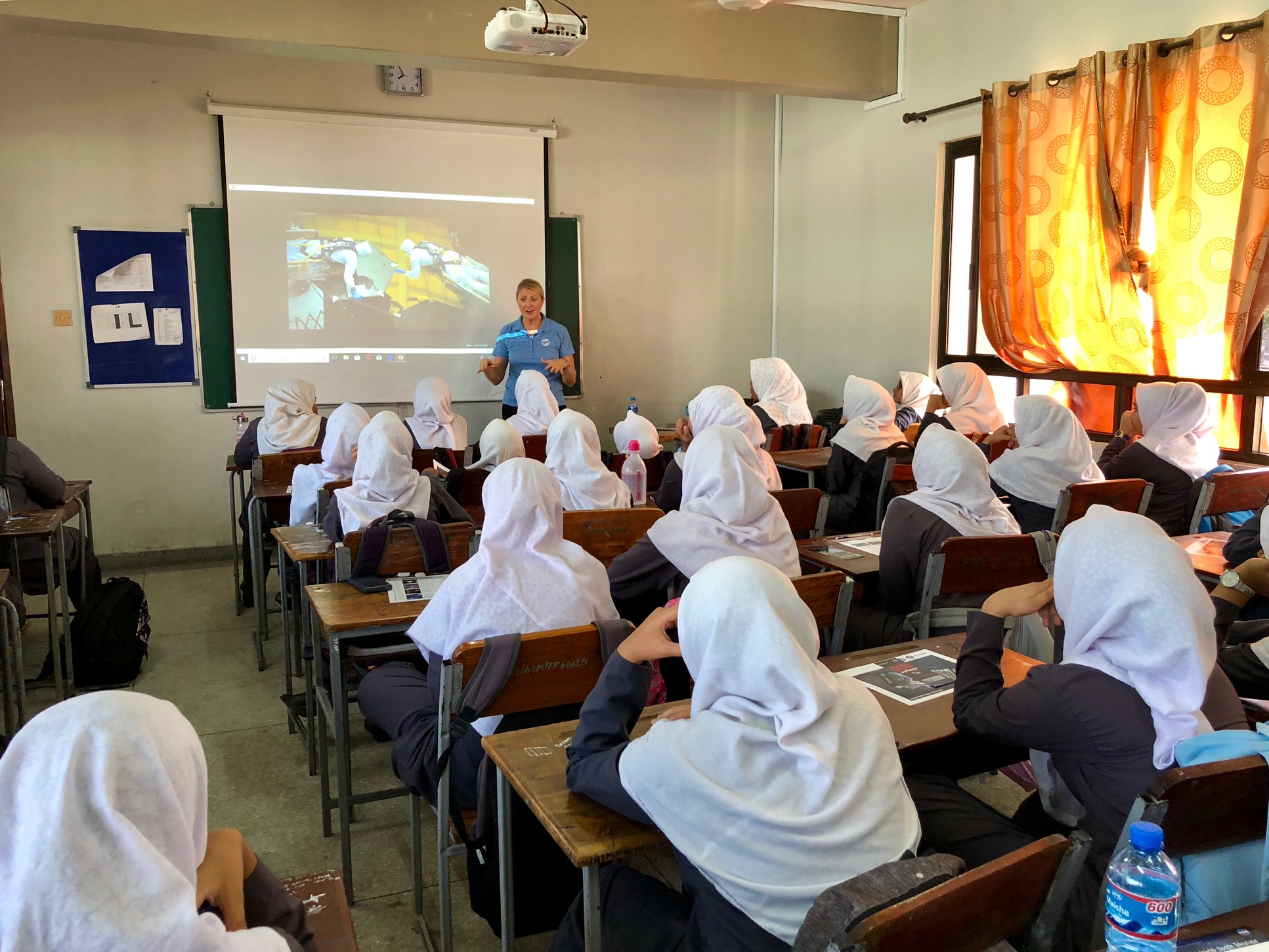 A woman with light skin and blonde hair wears a blue polo and stands in front of a classroom of about 20 grade 8 girls sitting in desks. The girls are wearing white hijabs and black long sleeve shirts.