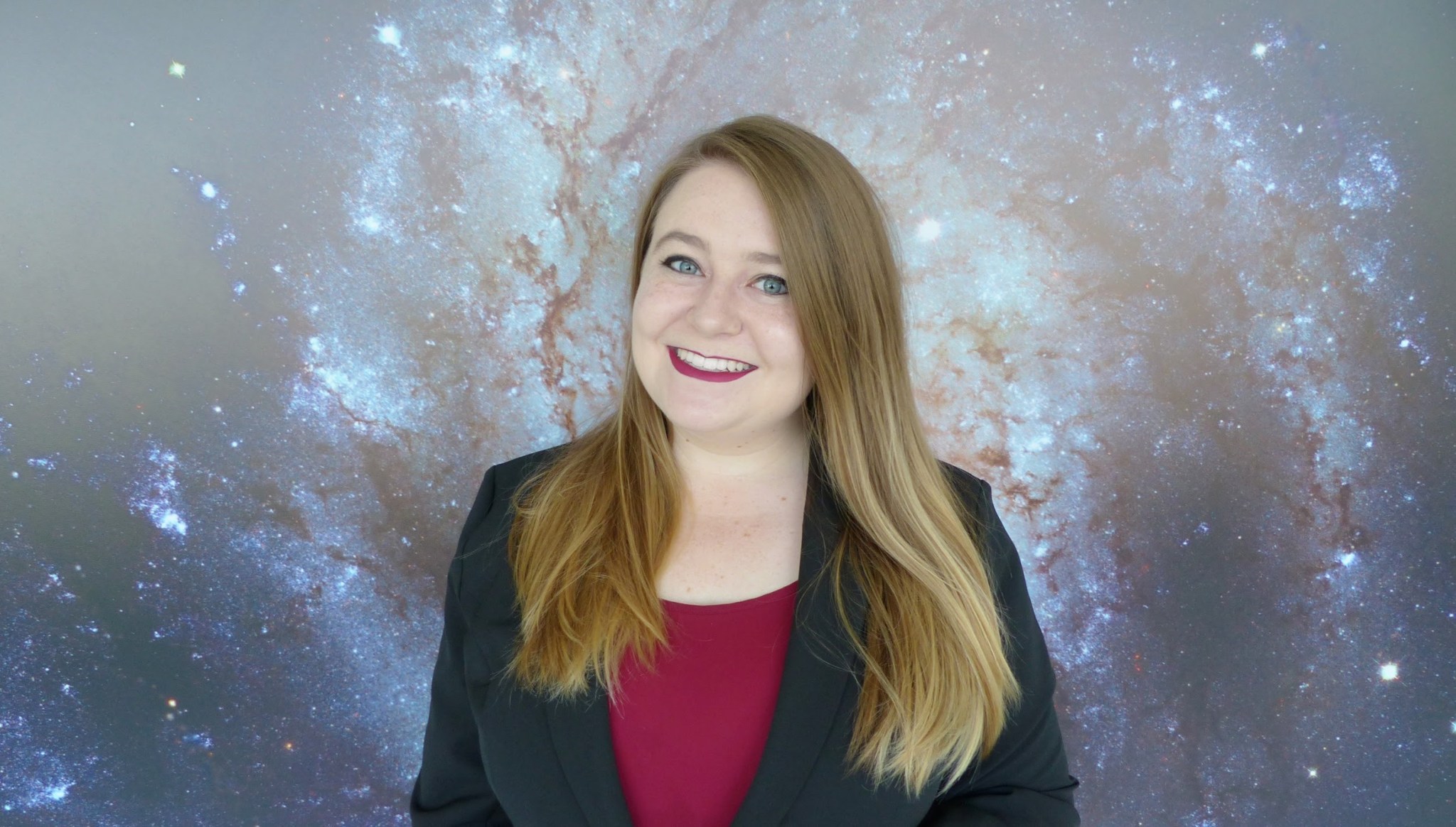 Woman with fair skin and shoulder length brown blonde hair wears a pink red shirt with a black blazer and marching red pink lipstick.