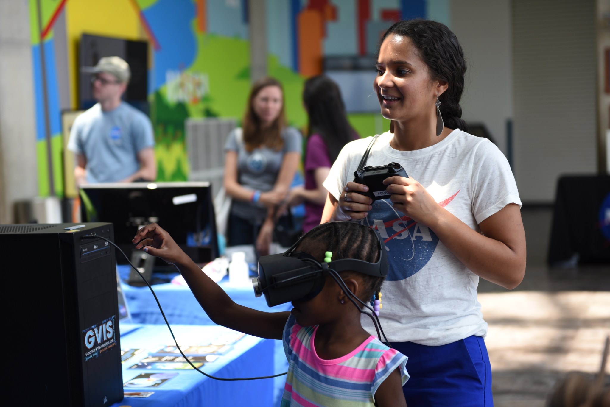 A woman wearing a white NASA t-shirt holds a gaming controller while standing next to a young girl who wears a virtual reality headset. 