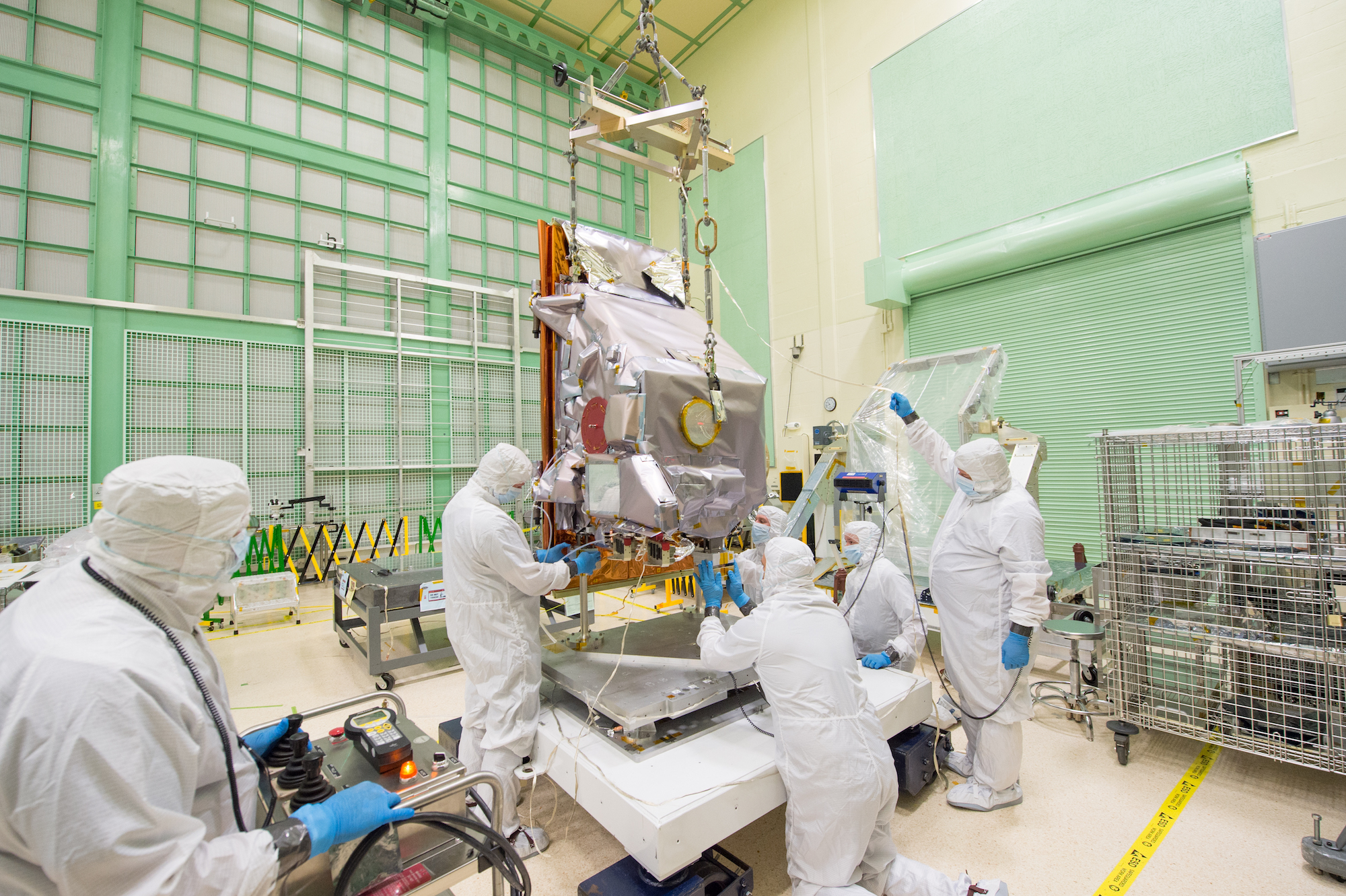 Engineers in white clean room suits with hoods and blue gloves and masks work on the TIRS-2 instrument inside a clean room with white and green walls. The instrument is wrapped in silver foil.