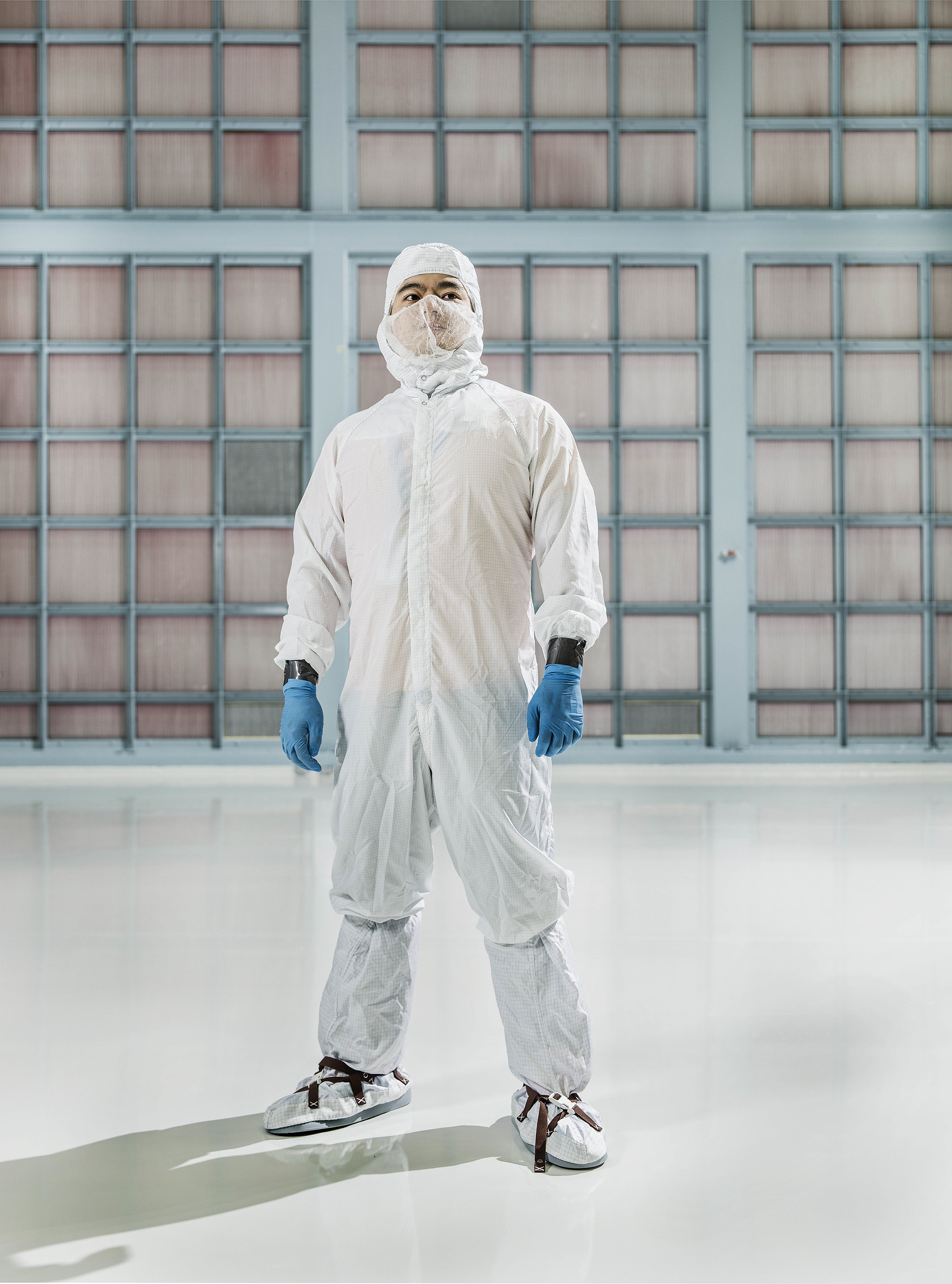 Man wears sterile "bunny suit," all white covering, including head, face, and feet. He wears blue gloves. He is standing in a cleanroom, in front of filters that go off frame the length of the wall behind him. 