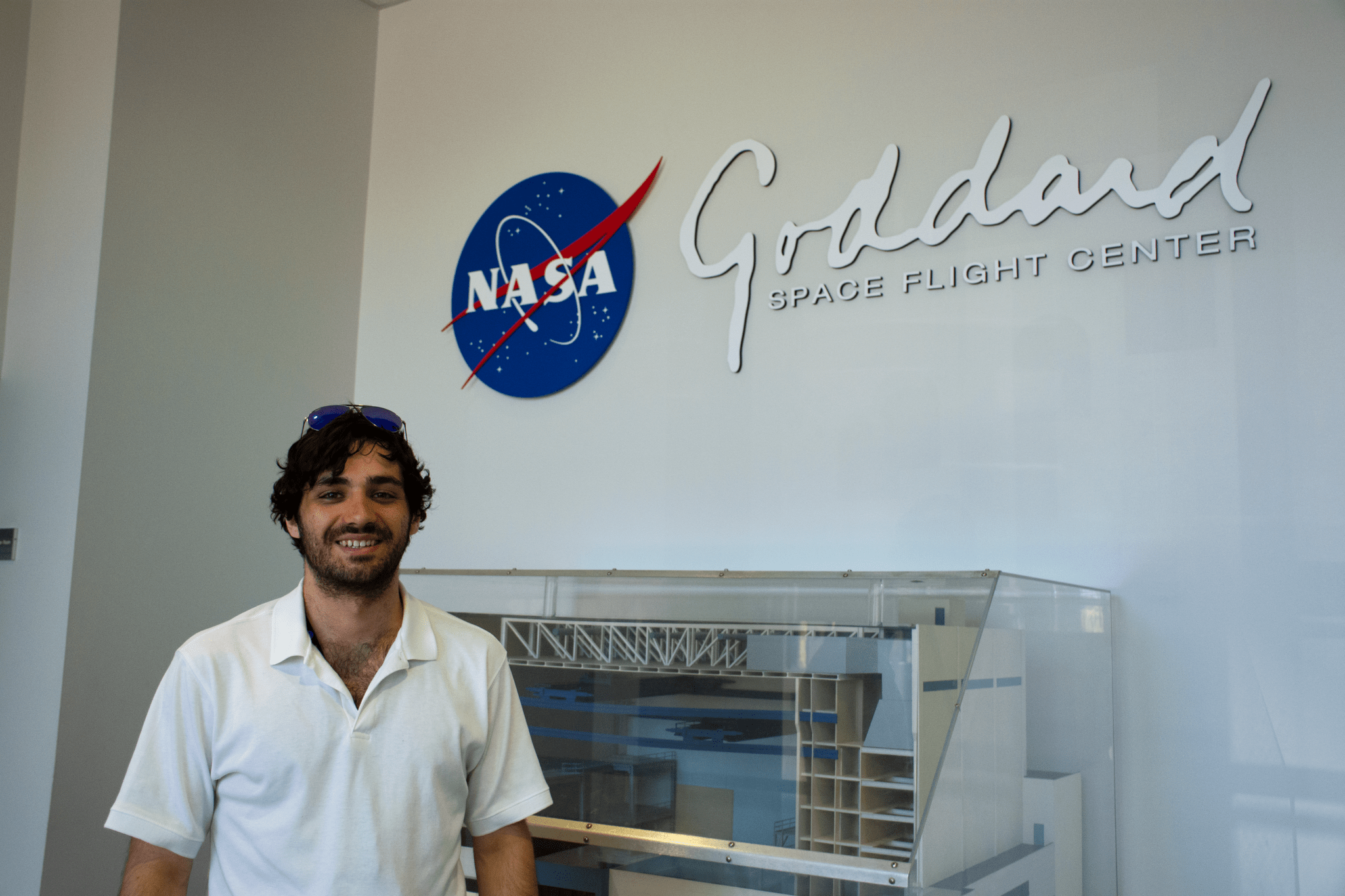 Man with black, curly hair wearing a white polo stands in front of a wall with the NASA Goddard logo.