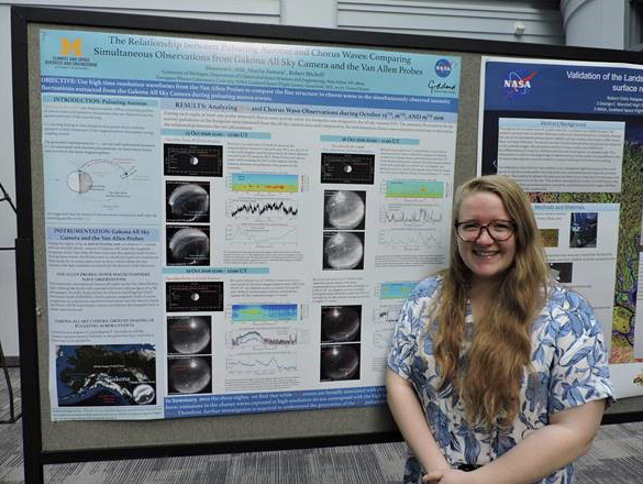 Woman with light skin and long blonde/brown hair and glasses wearing a blue floral shirt stands in front of a poster of her work 