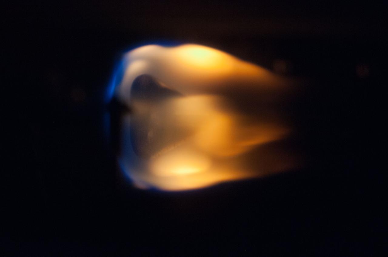 A nearly spherical flame points sideways, dark blue on its edge becoming bright yellowish orange in its center and darker orange at the end of several tongues of flame extending to the right.