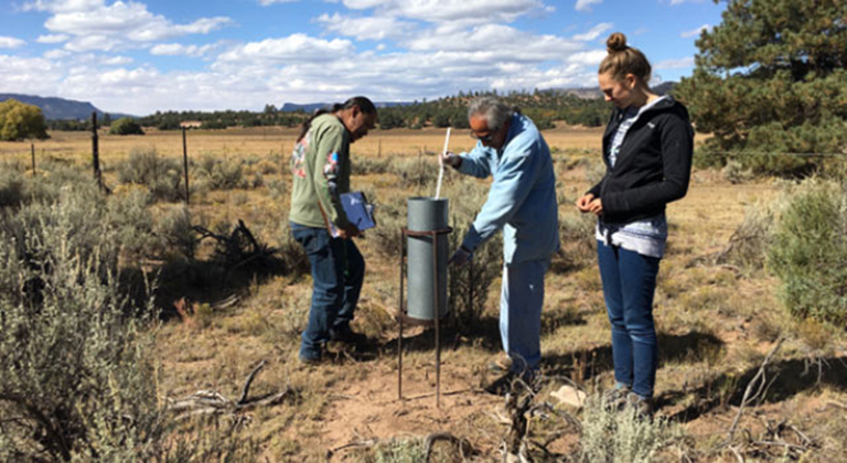 Three people stand in a desert with scrub brush around them. They are looking at a water container and taking measurements.