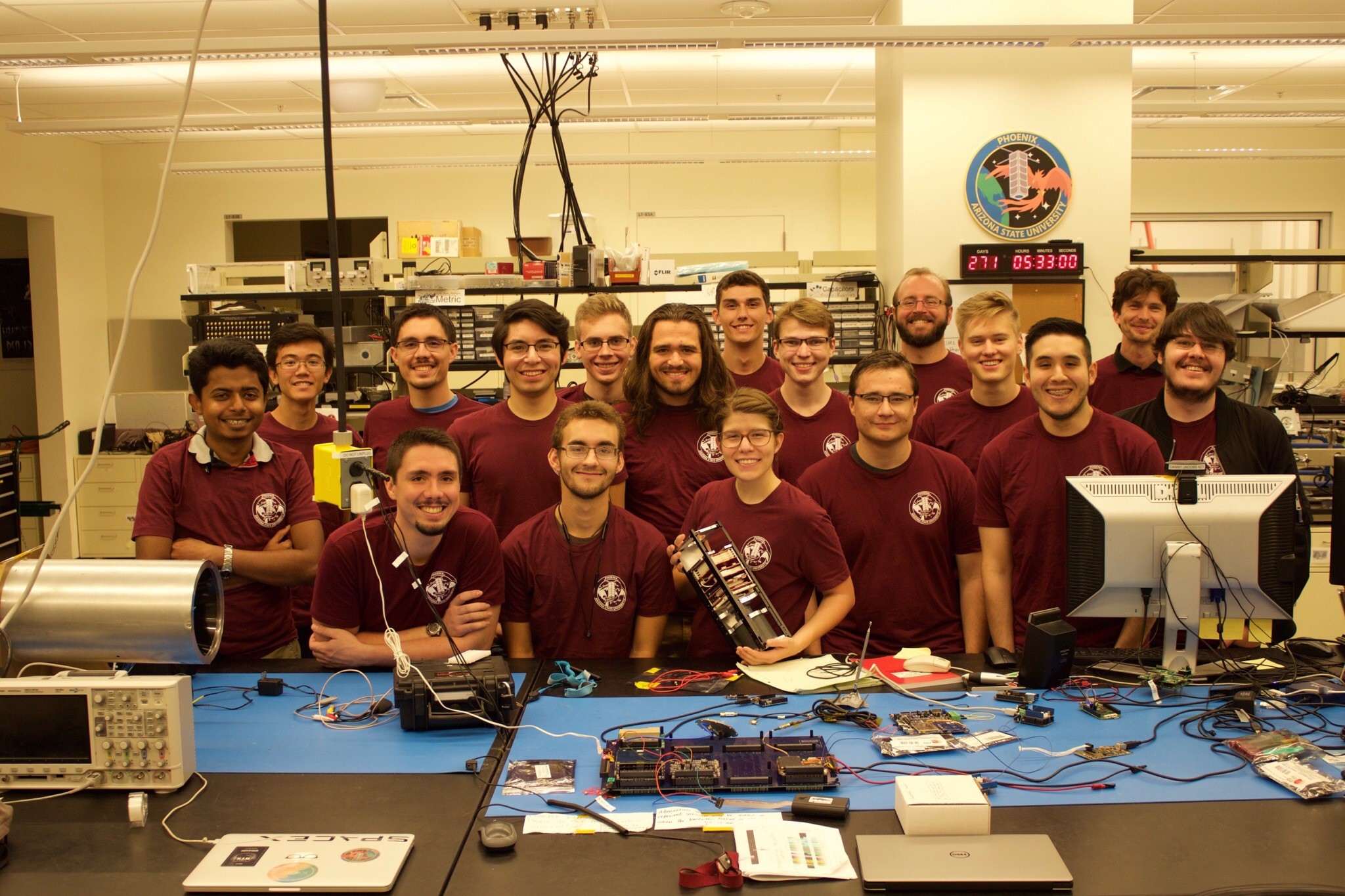 A group photo of multiple students all wearing maroon shirts. In front of them is a table with electronics on it.