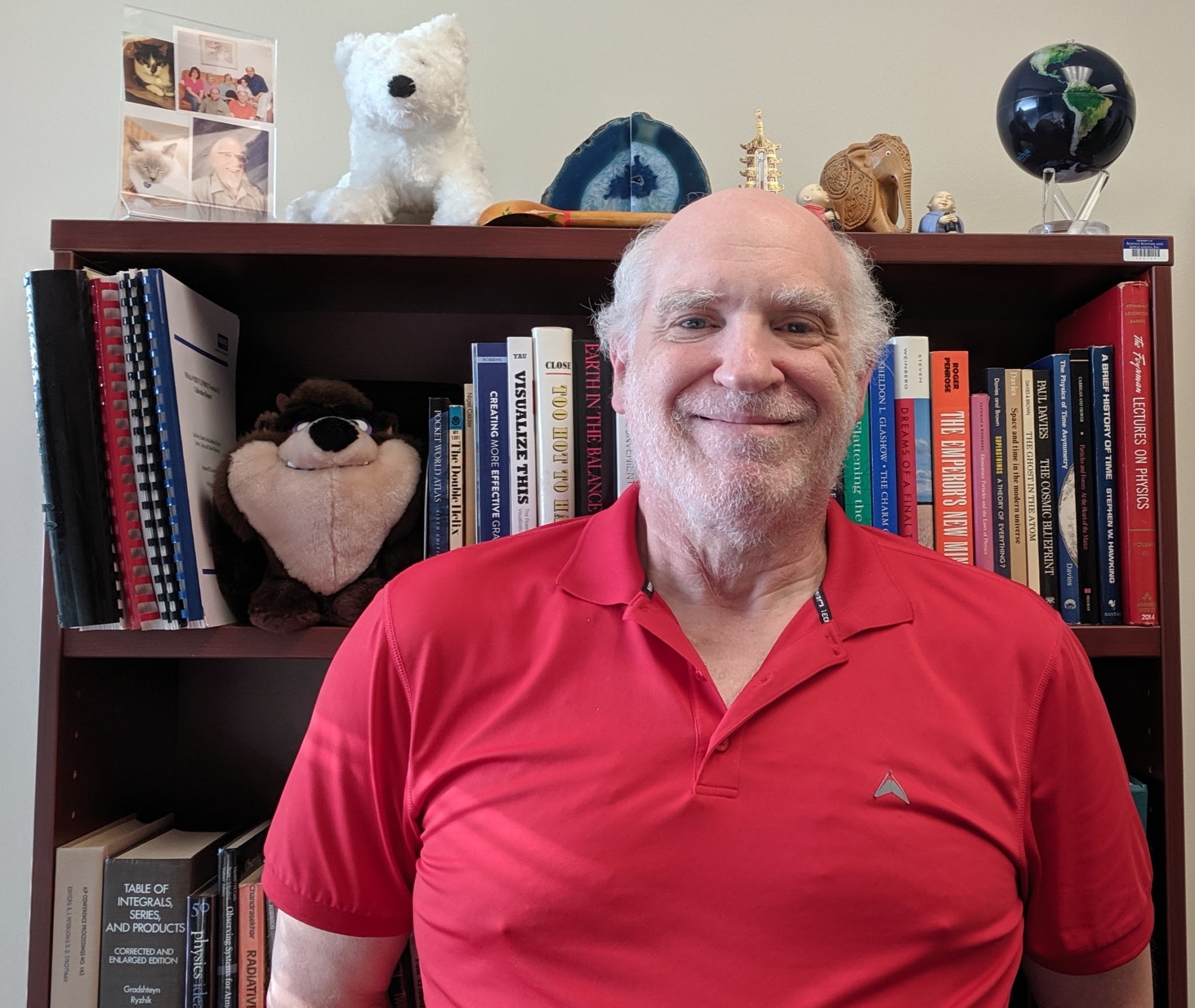 Man with fair skin and white hair wears a red polo shirt. He is smiling, standing in front of a bookcase. The bookcase has a variety of books and a stuffed Tasmanian Devil on the shelves. On the top of the book case, there are photos, a stuffed white dog, a globe, and other knick knacks.  