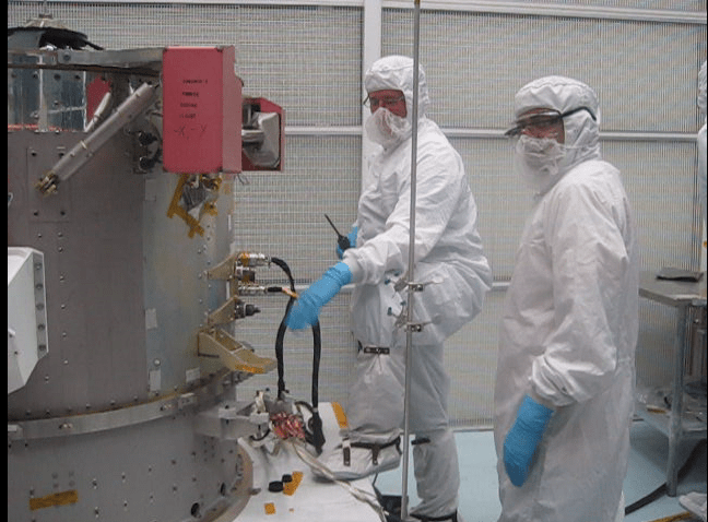 Two people wearing sterile, white "bunny suits" look at a cylinder machine with two hoses coming from it. 