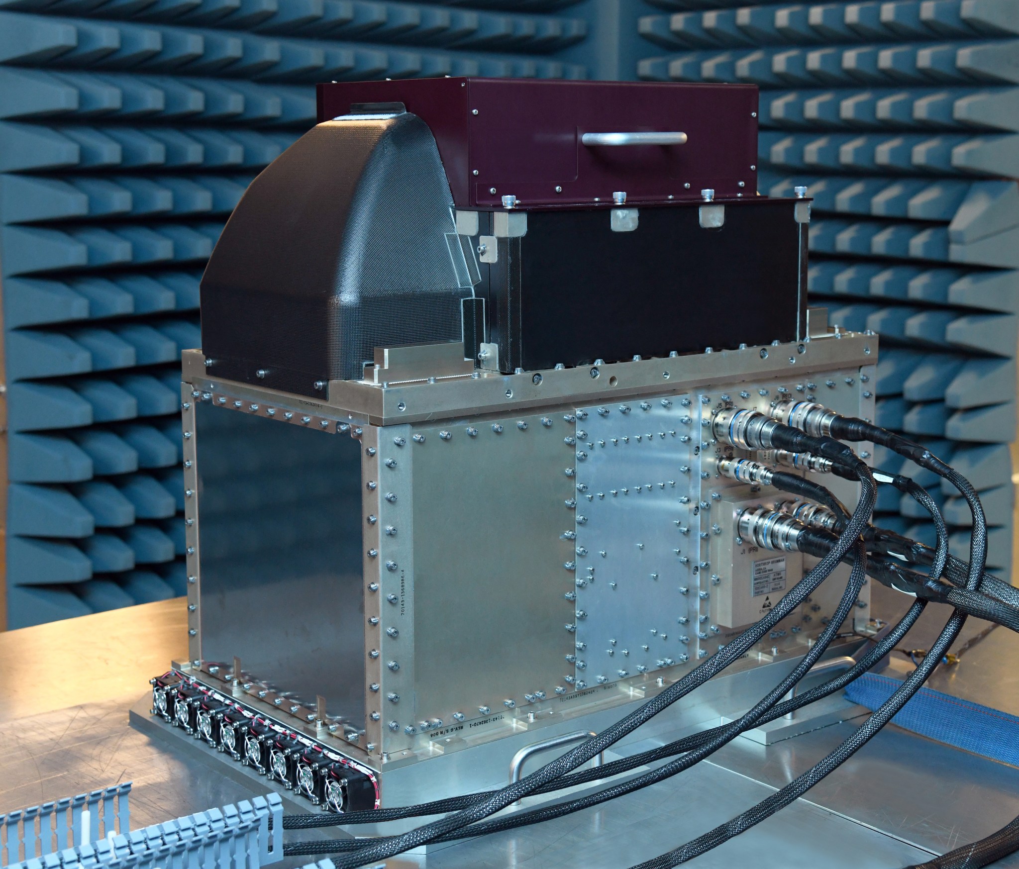 The Advanced Technology Microwave Sounder, or ATMS, an instrument shaped like a silver box with dark matte boxes stacked on top and cables plugged into the side. In this image, ATMS sits on a silver table in a room lined with foam spikes.