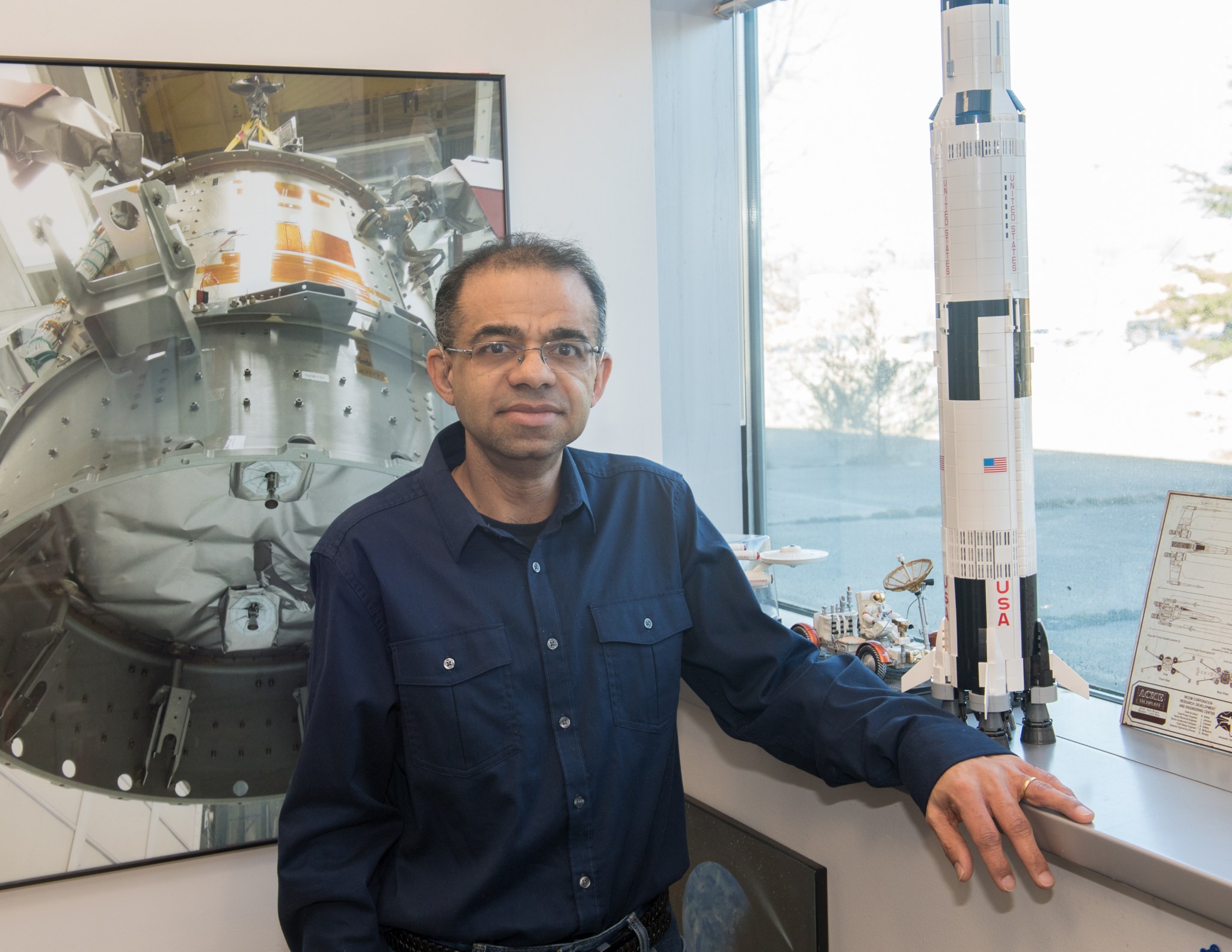 Man with dark tan skin wears glasses, a wedding ring, and a blue long sleeve shirt. He is standing next to a large photo of and a model of the Space Shuttle. 