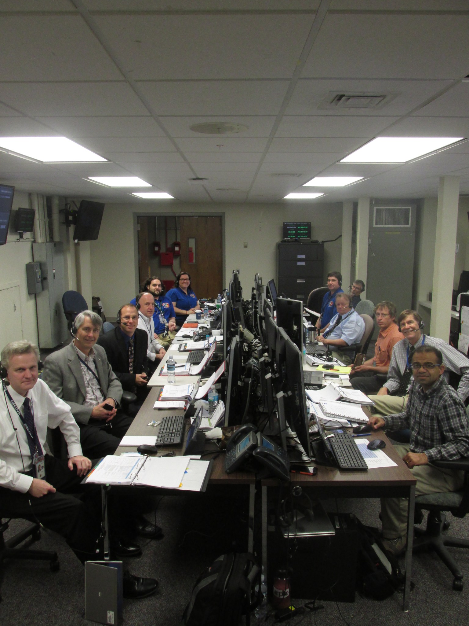 11 men and one woman sit on both sides of a table with computers and papers down the middle