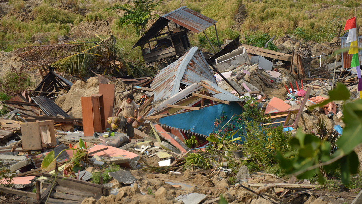 A man retrieving coconuts from a home destroyed by landslides, with damaged rice paddies in the background. 