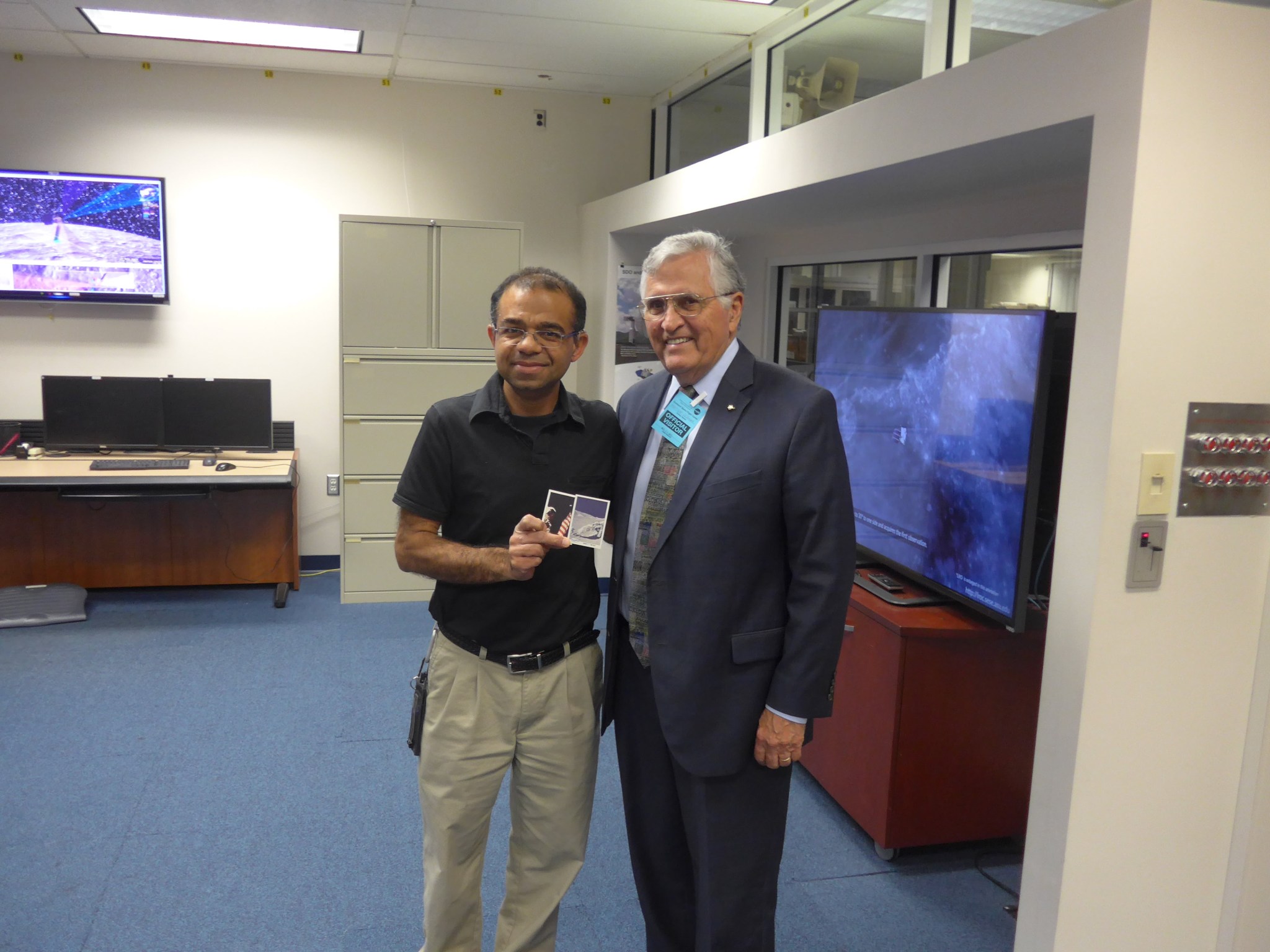 Two man stand together, smiling. The man on the left has dark tan skin, black hair, is wearing a black polo, tan pants, and is holding a post card. The man on the right has fair skin, white/grey hair and is wearing a navy suit and a blue "visitor" badge. They are standing in a conference room with blue carpet and two televisions on either wall. 