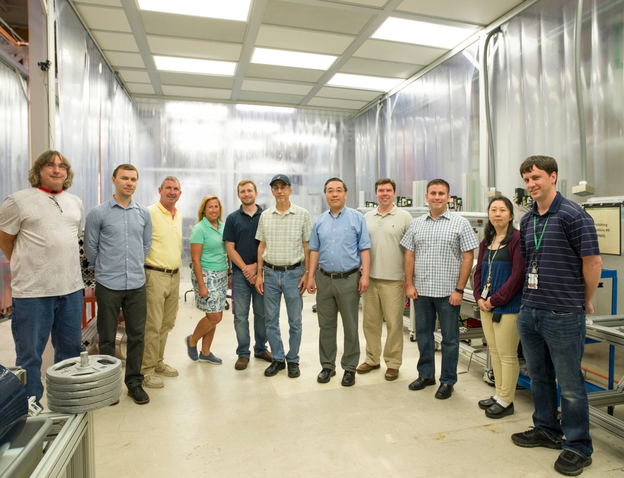 Group of 11 people, 9 men and 2 women standing side by side in a room with silver walls. 