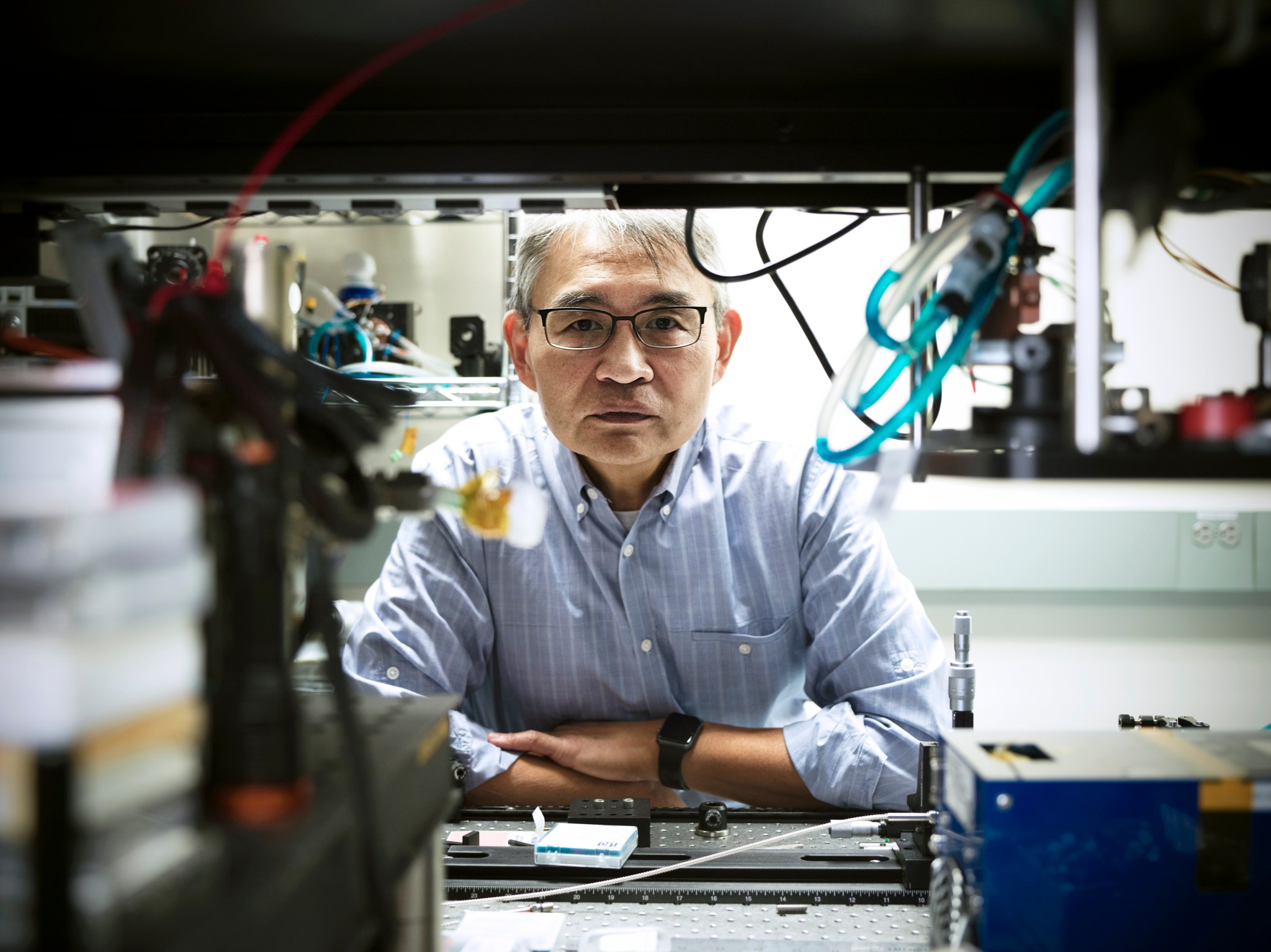 Goddard technologist Tony Yu, man with gray hair and tan skin, wearing glasses and a blue dress shirt sits and looks through a gap in wires and cords at his desk.