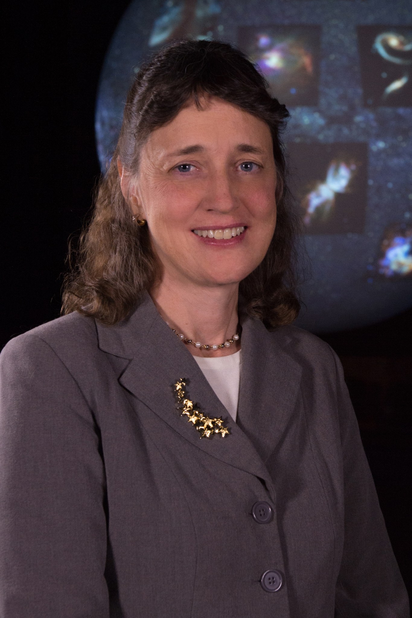 Woman with brown hair, pulled half up, wearing a grey blazer over a white shirt with a pearl necklace and gold broach with stars, smiles against a Hubble image background